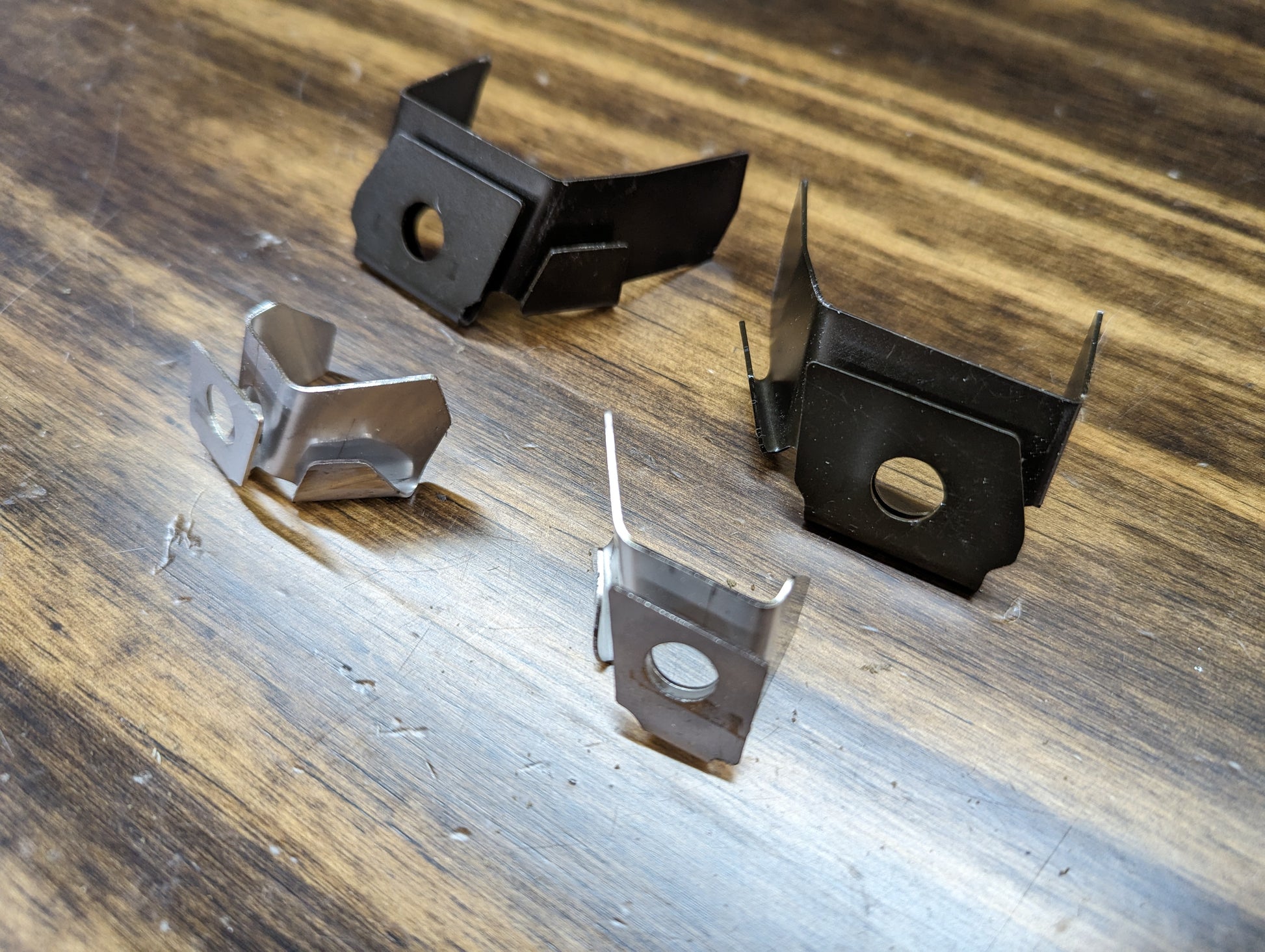 Four small metal brackets from The Ugly Truckling KIT Door Panel Armrest Repair for 1998-2005 Chevy S10, Blazer, GMC Sonoma, and Jimmy are shown on wood—two black and two silver, each with bent tabs and angled shapes.