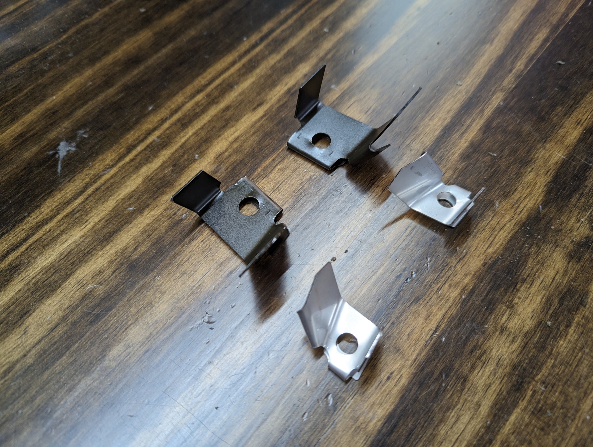 Four small, metallic clips from The Ugly Truckling KIT Door Panel Armrest Repair for 1998-2005 Chevy S10, Blazer, GMC Sonoma, and Jimmy are arranged on a wooden surface, showing slight variations in shape and orientation.