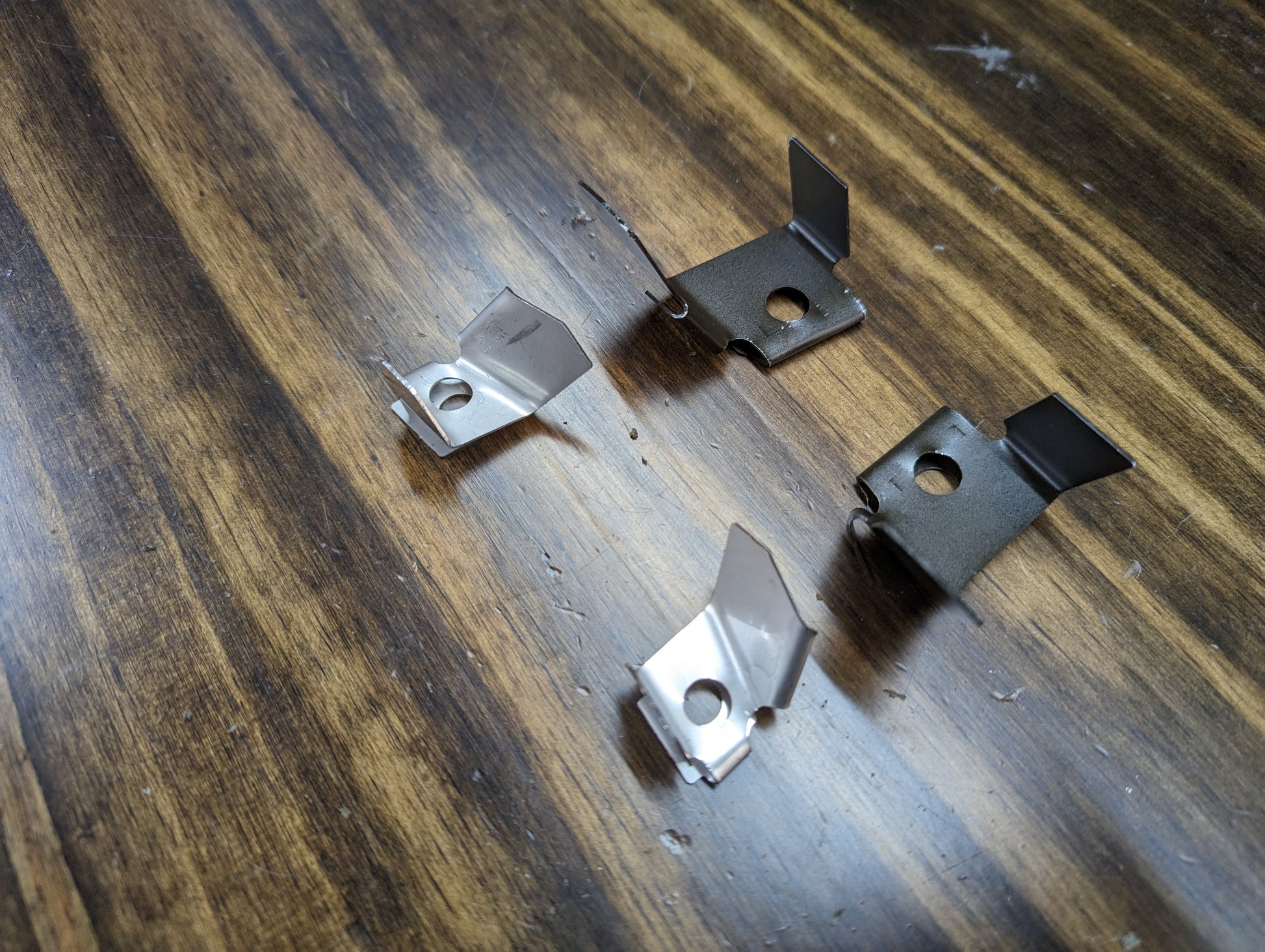 Four small metal brackets from The Ugly Truckling KIT Door Panel Armrest Repair for 1998–2005 Chevy S10, Blazer, GMC Sonoma, and Jimmy are arranged on a wooden surface with metal shavings nearby.