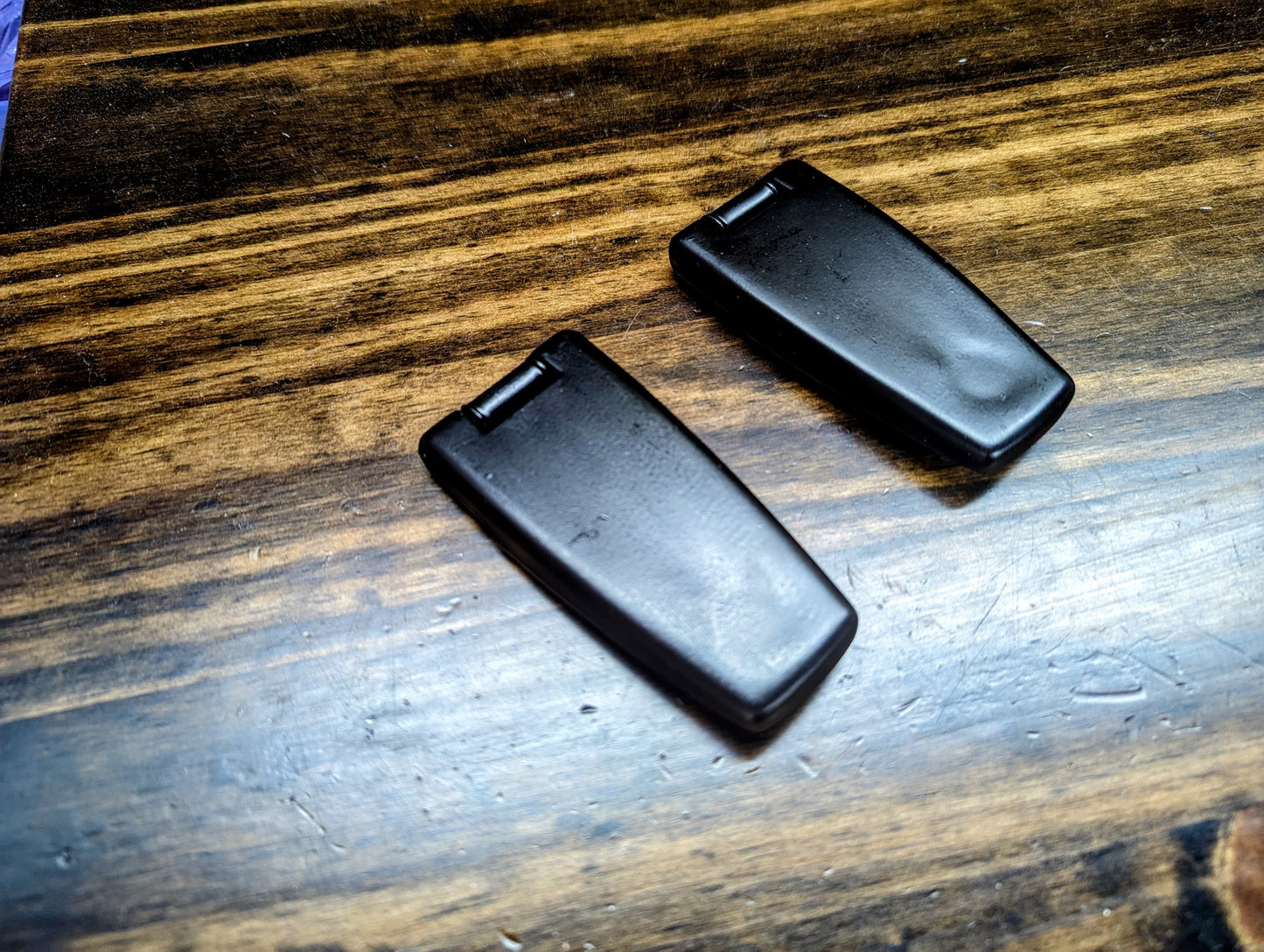 Two black, rectangular General Motors OEM Rear Hatch Glass Hinges for 1983-1994 Chevrolet S10 Blazer are shown side by side on a wooden surface with visible grain.