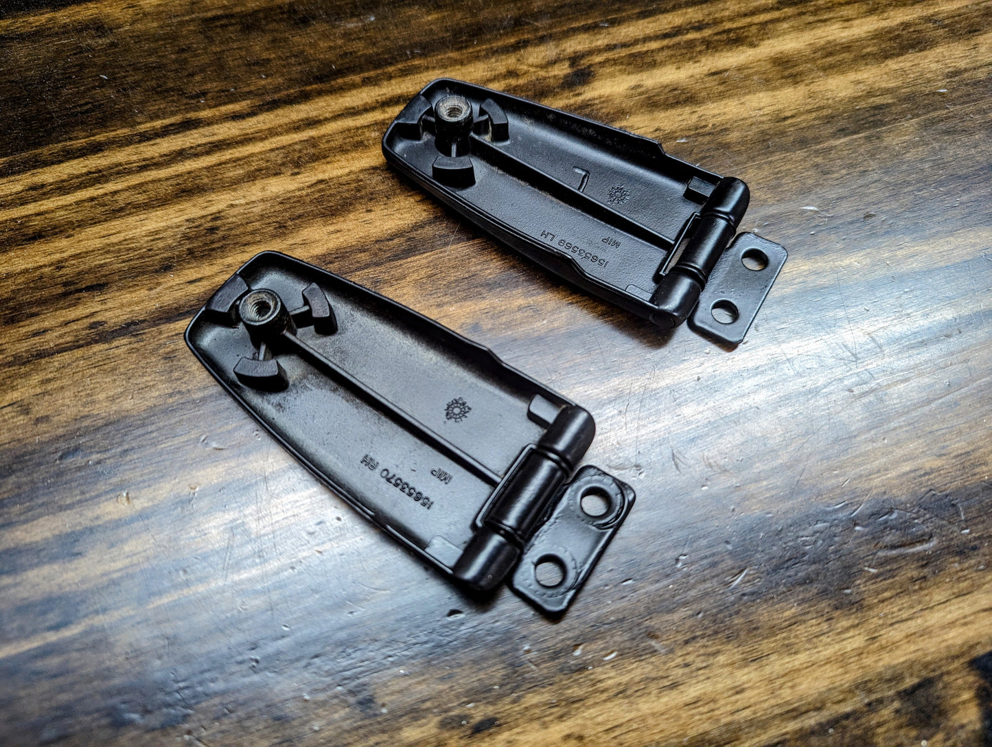 Two General Motors OEM Rear Hatch Glass Hinges for 1983-1994 Chevrolet S10 Blazer and more are shown on a wooden surface, each with mounting plates, visible screws, and markings, positioned diagonally to each other.