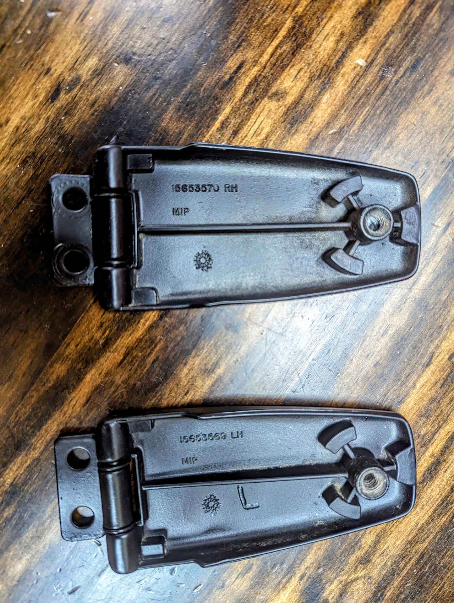 Two black OEM Rear Hatch Glass Hinges for 1983-1994 Chevrolet S10 Blazer by General Motors, marked RH and LH with molded serial numbers and icons, rest on a wooden surface.