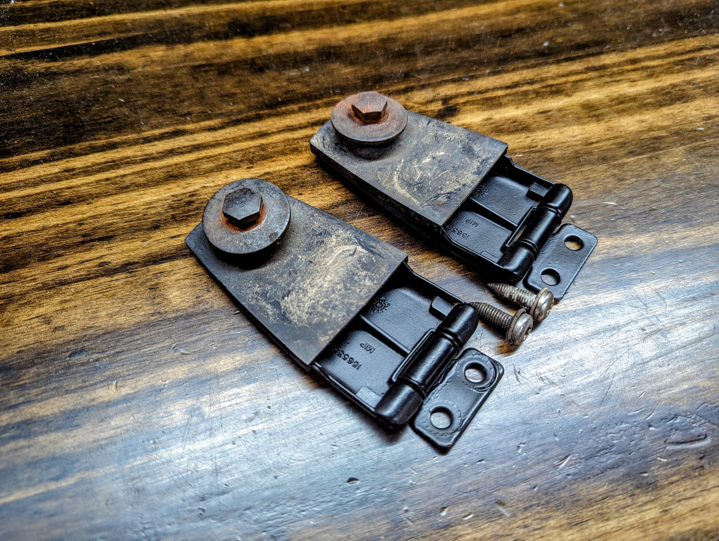 Two OEM Rear Hatch Glass Hinges for 1983-1994 Chevrolet S10 Blazer by General Motors, with large bolts attached, are lying on a wooden surface and show some wear and rust.