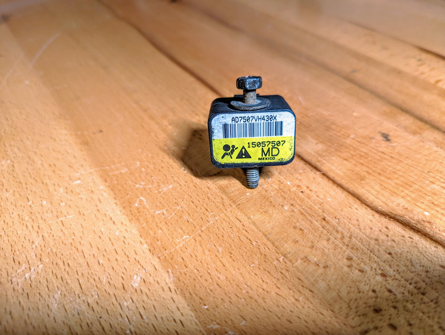 A General Motors TESTED Front Impact Air Bag Sensor (Part Number 15057507) for 2003-2004 GM Trucks rests on a wooden surface, displaying a yellow warning symbol on its label.