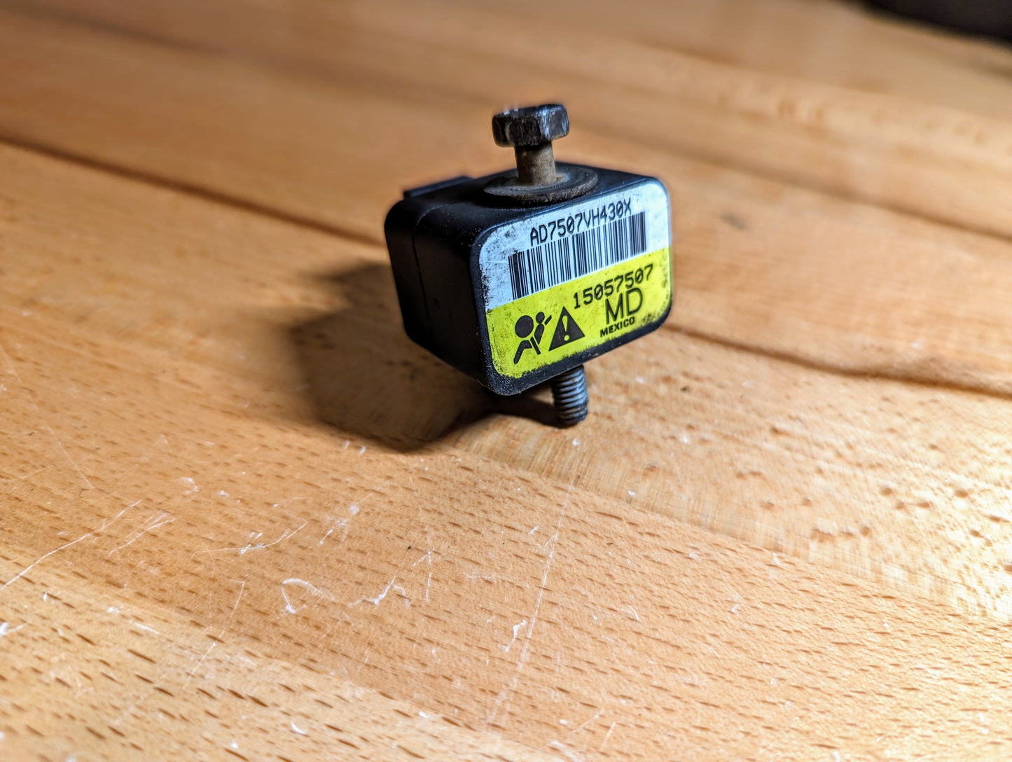 A General Motors TESTED Front Impact Air Bag Sensor (Part #15057507) for 2003-2004 GM Trucks, featuring a yellow warning label, barcode, and screw, rests on a wood-grain surface.