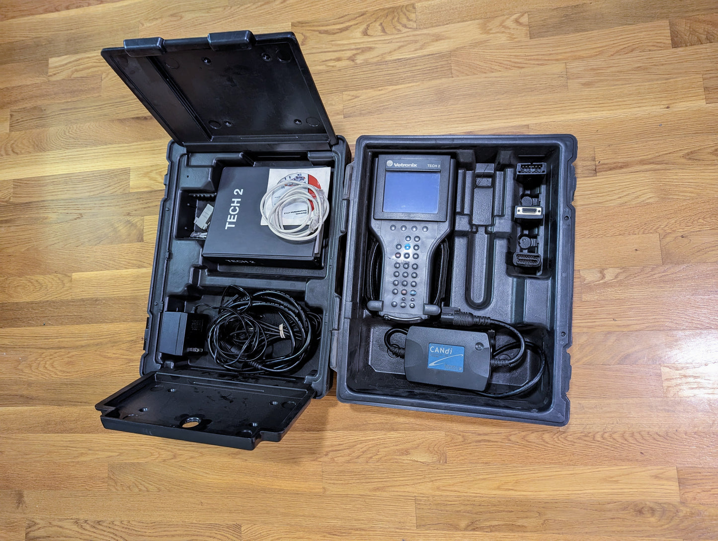 A black hard case contains the Vetronix TESTED Tech 2 Automotive Diagnostic Tool & Accessories for 1991-2013 GM vehicles, including cables, manuals, and a handheld OBD2 scanner, all neatly displayed on a wooden floor.