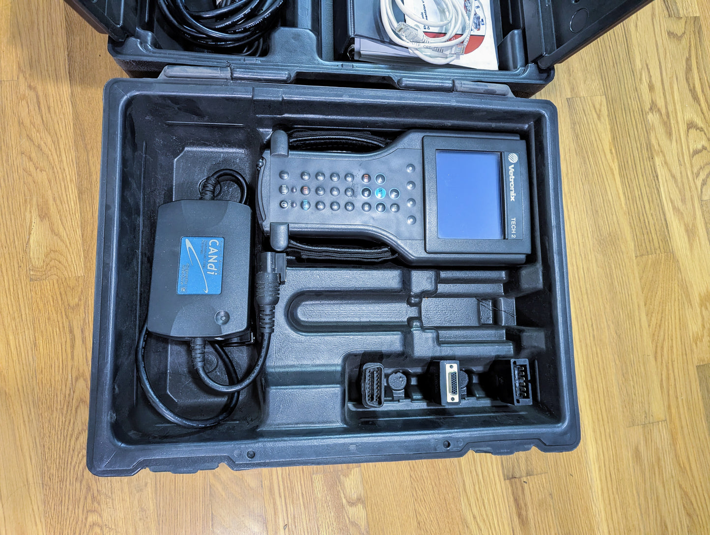 A black plastic case with a Vetronix TESTED Tech 2 Automotive Diagnostic Tool for 1991-2013 GM vehicles, power adapter, and accessories—neatly organized in custom compartments on a wooden floor.