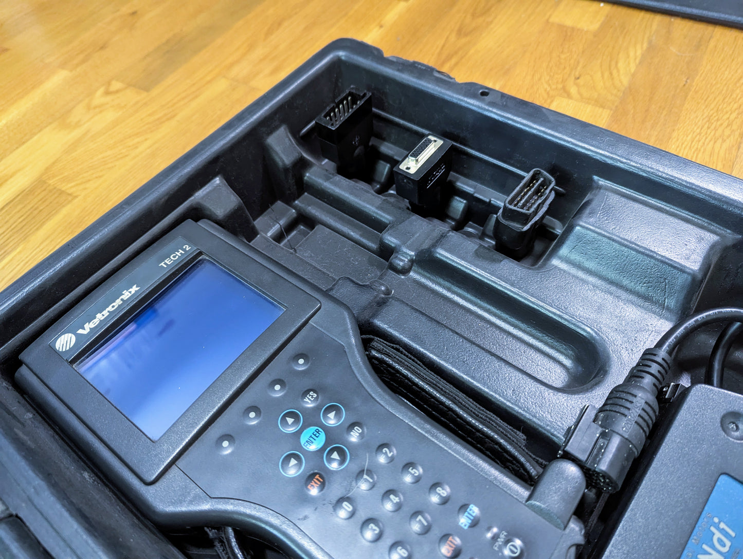A Vetronix TESTED Tech 2 Automotive Diagnostic Tool & Accessories for 1991-2013 GM Vehicles in a black case, shown with connector plugs and handheld device, on a wooden surface.