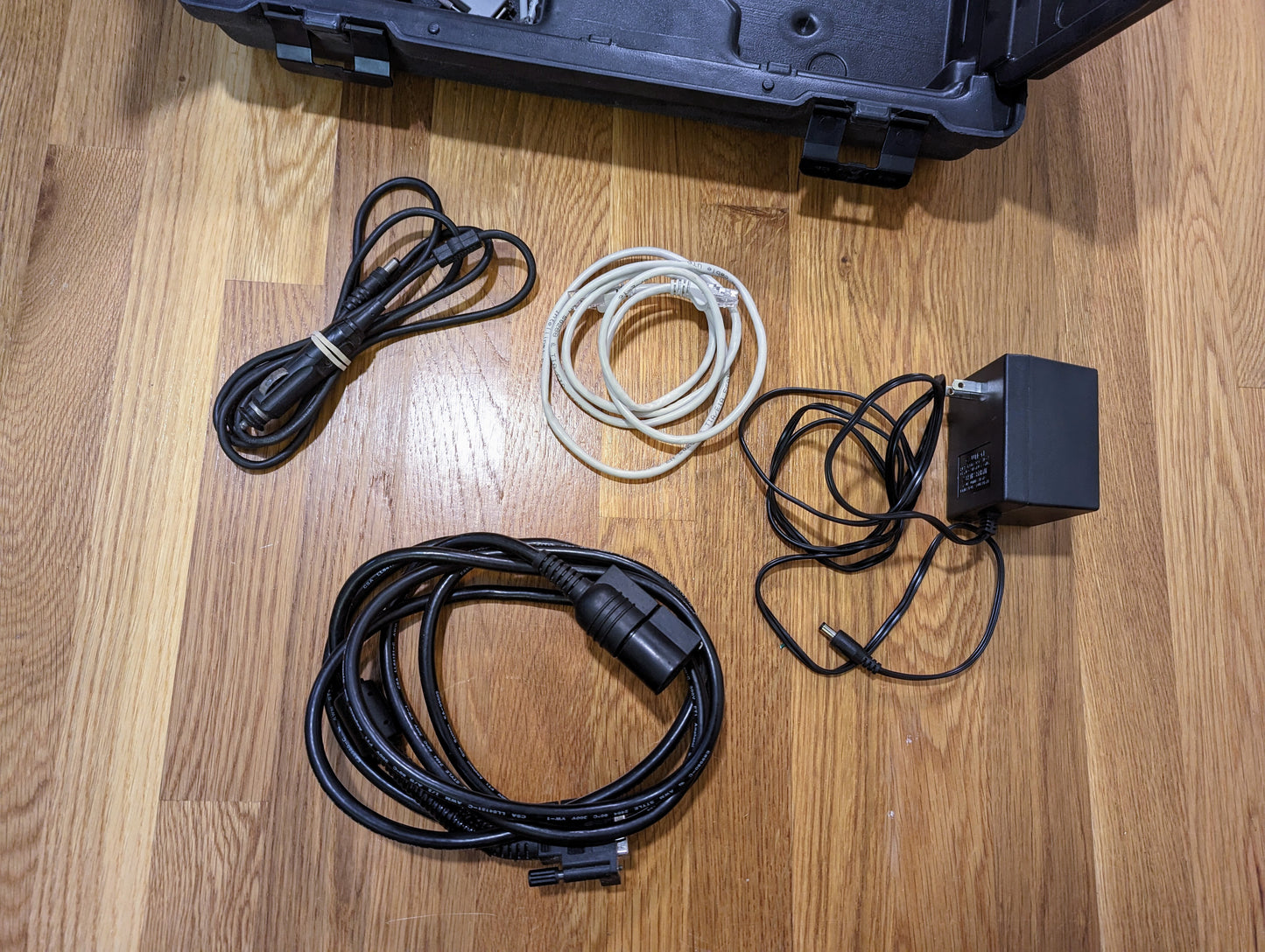 Four cables—a black power cord, a white Ethernet cable, a thin black USB cable, and a black power adapter—are arranged on a wooden floor beside an open Vetronix Tech 2 Automotive Diagnostic Tool & Accessories case for GM vehicles (1991-2013).