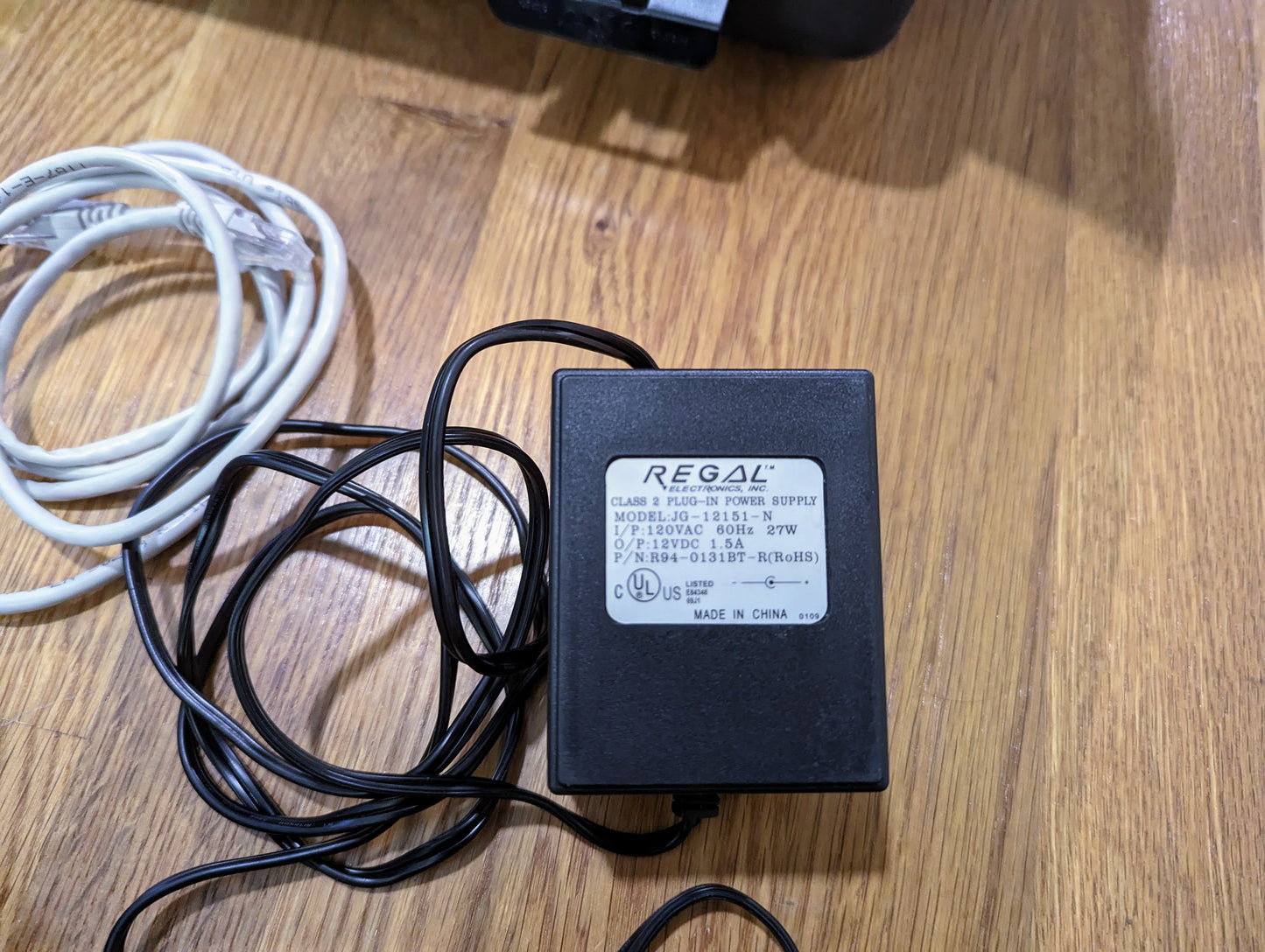 A black Vetronix power supply adapter with white label, model 18-011-03R, rests on wood near tangled cables—perfect for powering the TESTED Tech 2 Automotive Diagnostic Tool & Accessories for 1991-2013 GM vehicles.