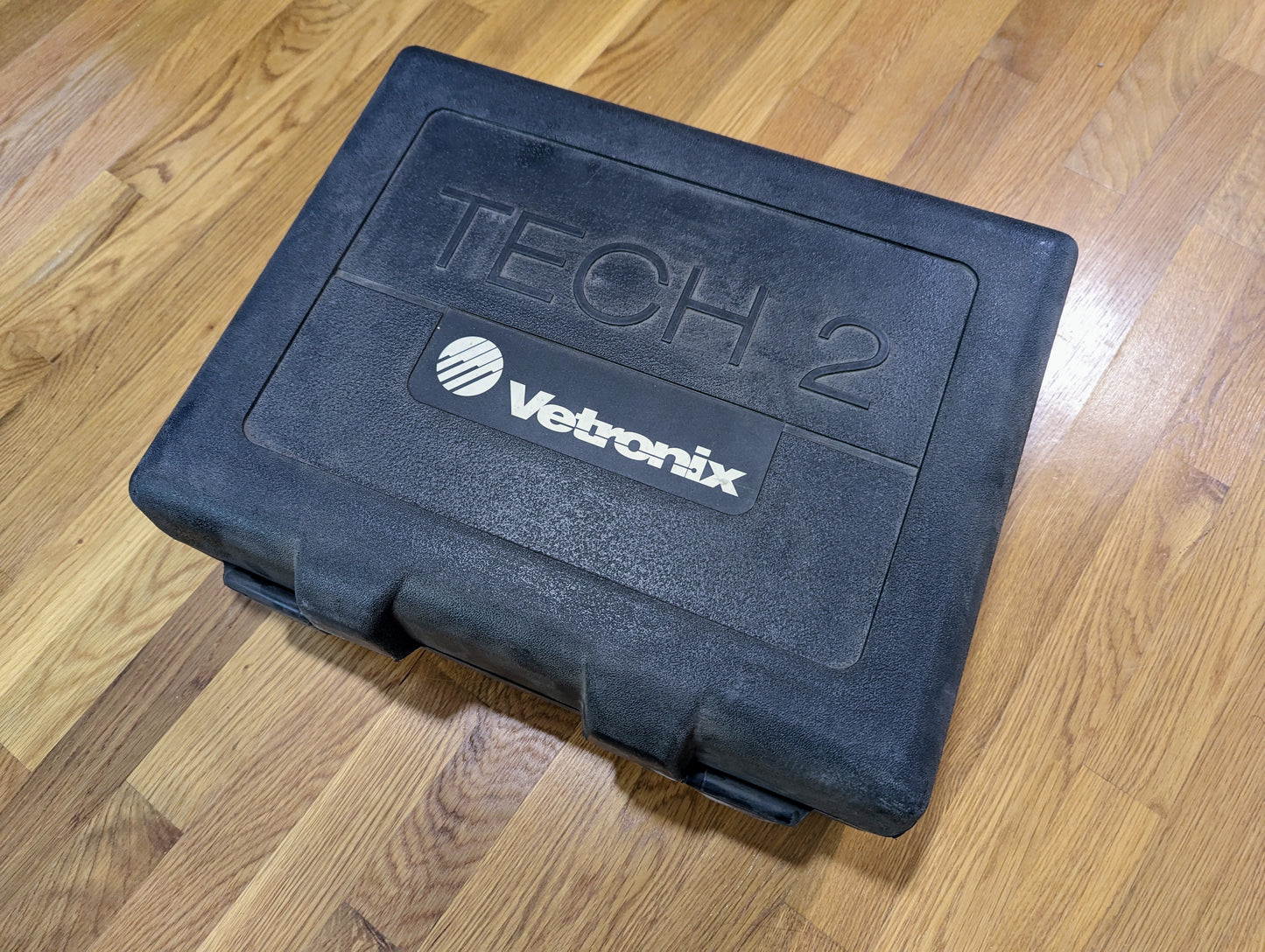 A closed black plastic carrying case labeled TECH 2 and Vetronix, containing a TESTED Tech 2 Automotive Diagnostic Tool & Accessories for 1991-2013 GM Vehicles by Vetronix, sits on a wooden floor.