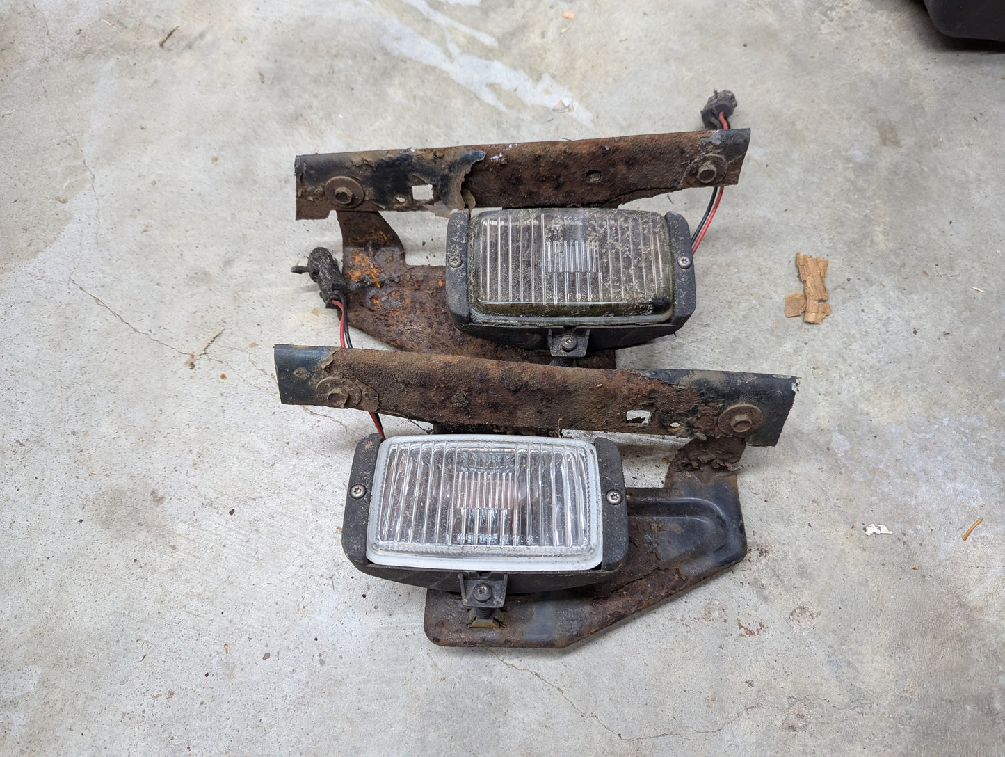A pair of OEM General Motors fog light assemblies with brackets for 1991-1994 Chevy S10 and Blazer lie on a concrete floor, showing significant corrosion and wear, with exposed wires and visible rust on the metal mounting brackets.