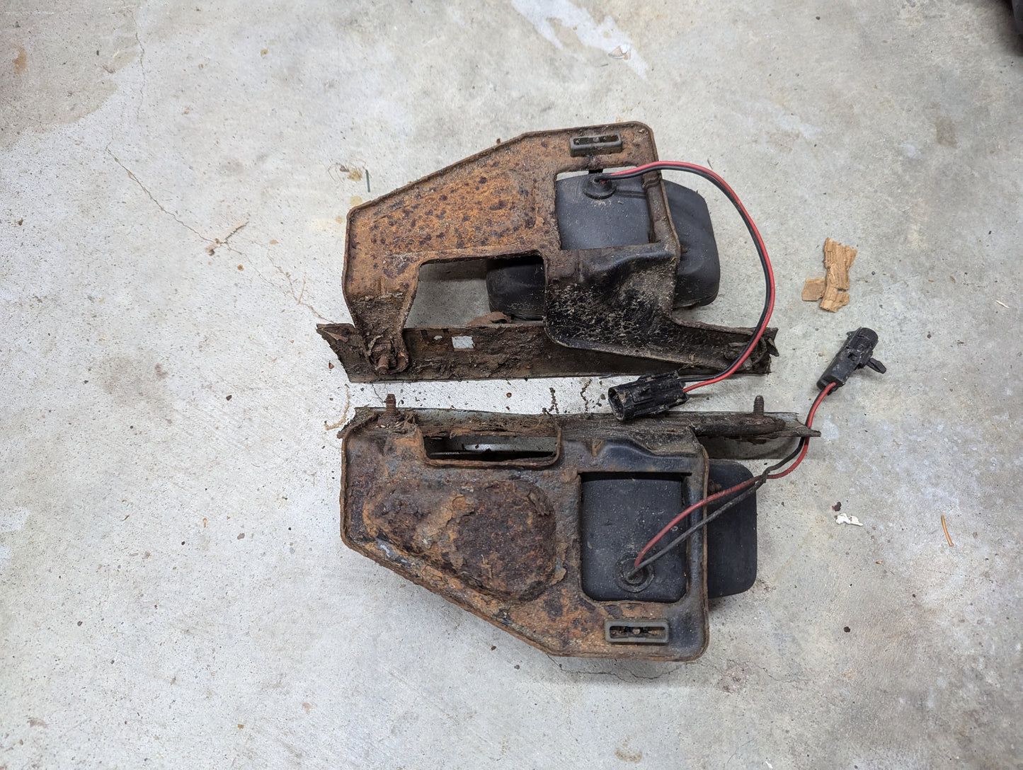 Two rectangular metal objects with black plastic parts and red/black wires on concrete—likely a General Motors OEM pair of fog light lamp lens assemblies with brackets for 1991-1994 Chevy S10, Blazer, and more.