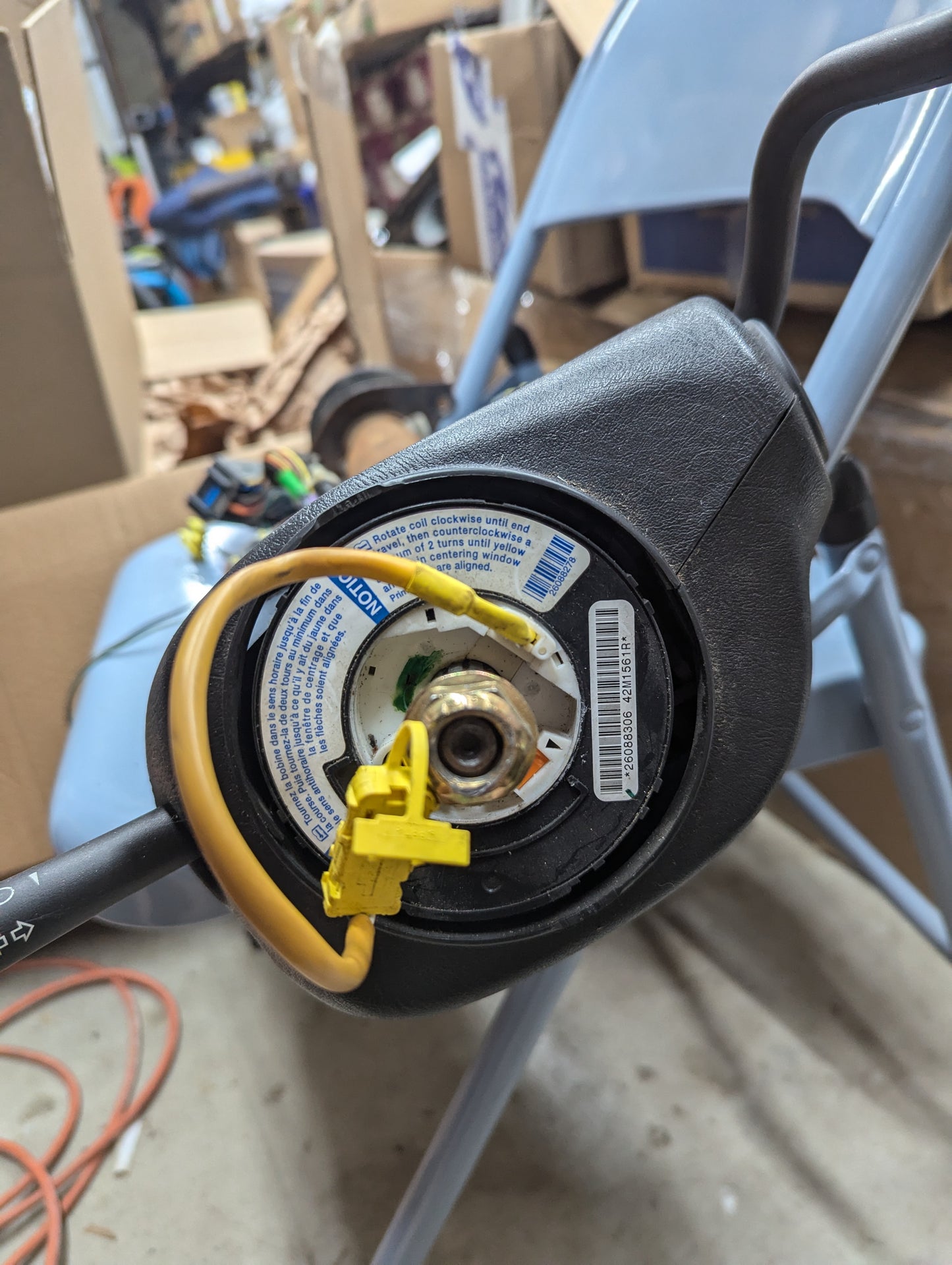 Close-up of a General Motors TESTED Steering Wheel Air Bag SRS Clock Spring OEM for 2000-2003 Chevy S10 Blazer, surrounded by cardboard boxes and tools in a cluttered garage setting.