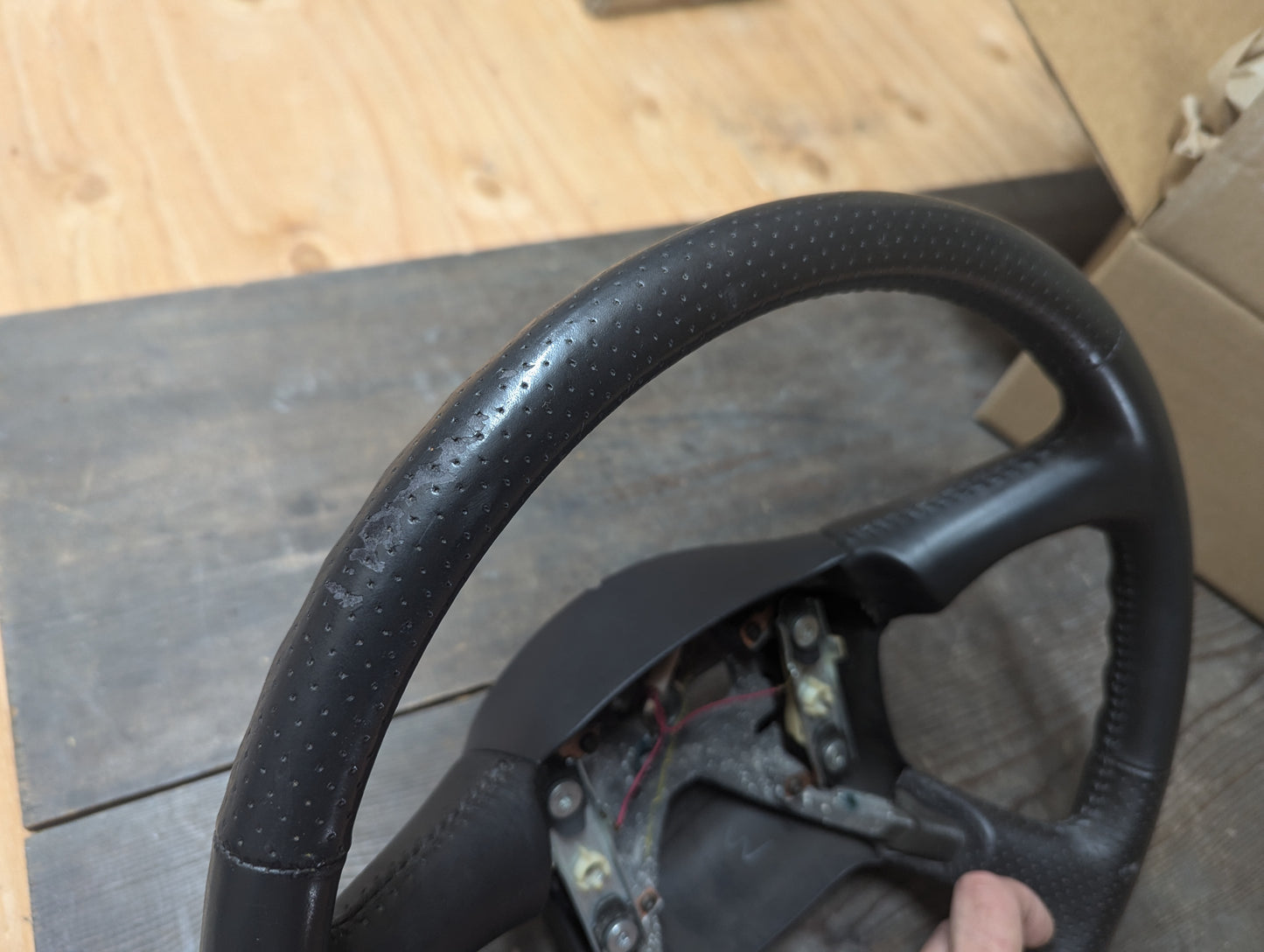 A close-up of a General Motors OEM Perforated Leather Steering Wheel for 1999-2005 Chevy Silverado, S10, and more shows visible wear, with a wooden surface and partially open cardboard box in the background.