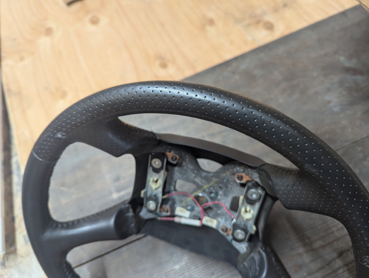 A close-up of a General Motors OEM Perforated Leather Steering Wheel for 1999-2005 GM Chevy Silverado and S10, partially disassembled to show mounting points and wiring, with textured perforated leather on a wooden surface.