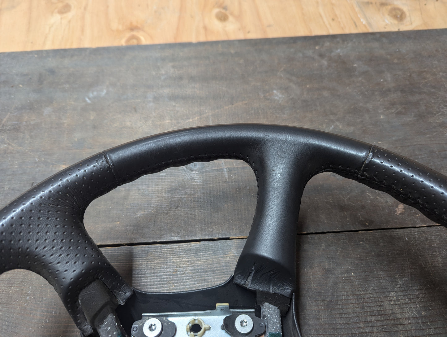 Close-up of a General Motors OEM perforated leather steering wheel for 1999-2005 GM Chevy Silverado S10, featuring detailed stitching and textured grip, shown on a dark wood surface with a lighter wood background above.