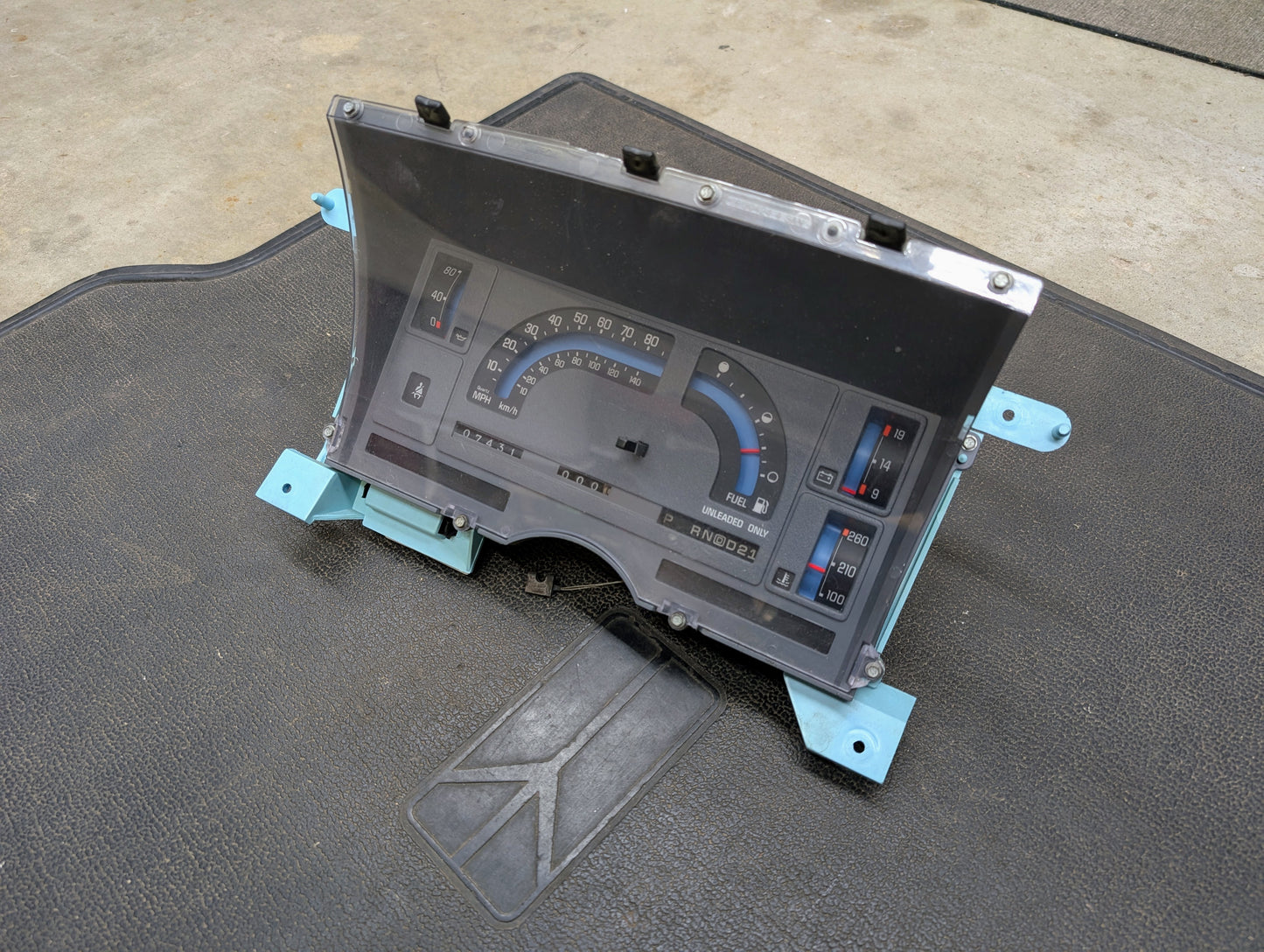 The Low Mile 74K OEM Instrument Cluster with PRNDL by General Motors for 1986-1994 Chevy S10 Blazer sits on a textured black mat atop concrete, displaying speed, fuel, and indicators through its clear digital-analog display cover.