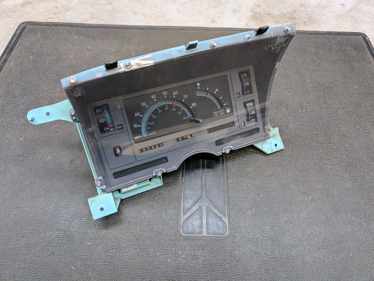 A General Motors Low Mile 107K OEM Instrument Cluster for 1986-1994 Chevy S10, Blazer, and more with a clear cover is shown on a black mat. This analog display provides speed, fuel level, and key readings from the original era.