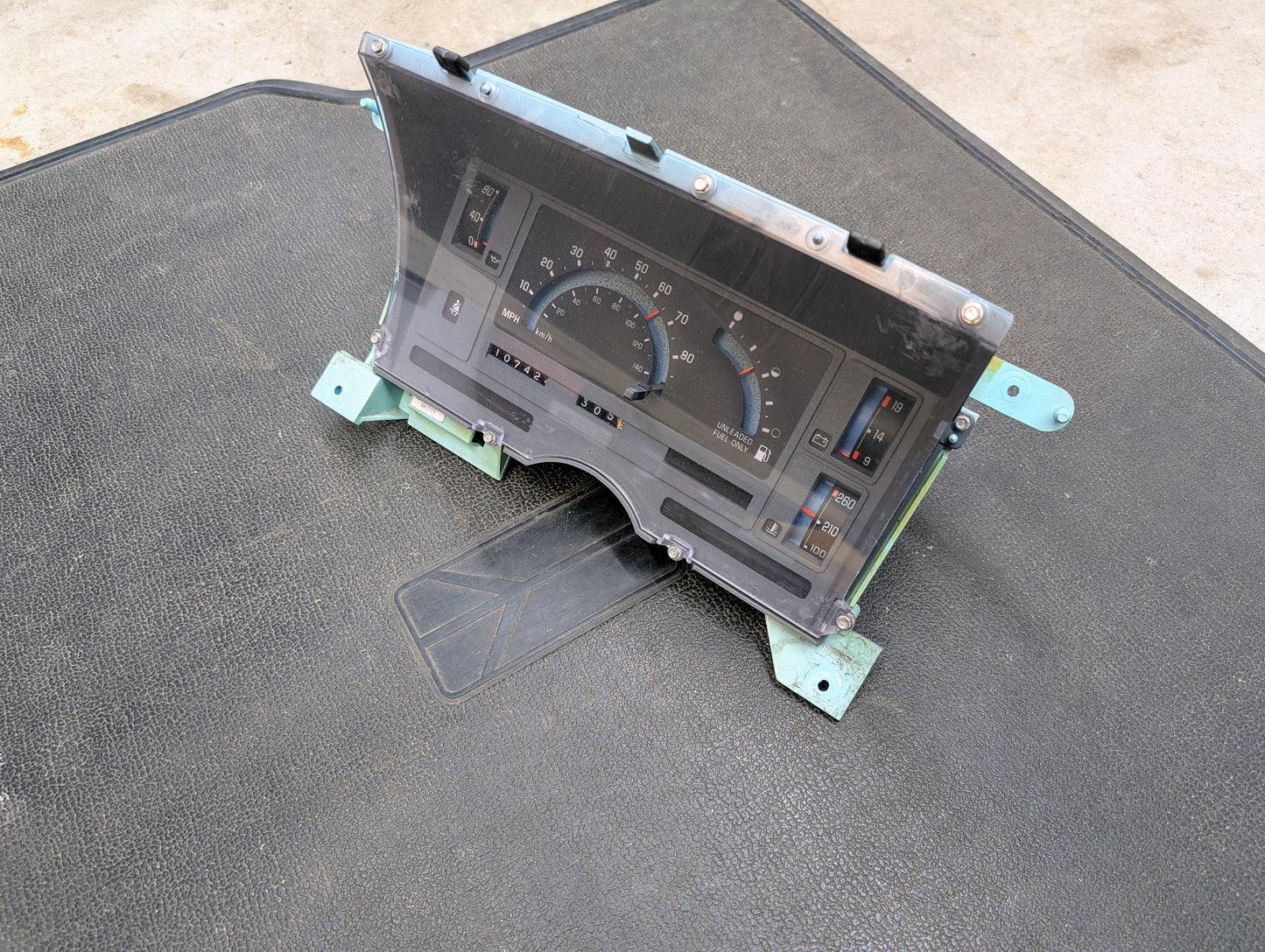 A Low Mile 107K OEM Instrument Cluster for 1986-1994 Chevy S10 Blazer by General Motors is displayed on a textured black mat, showing its speedometer, fuel and temperature gauges, with metal mounting brackets visible at the sides.
