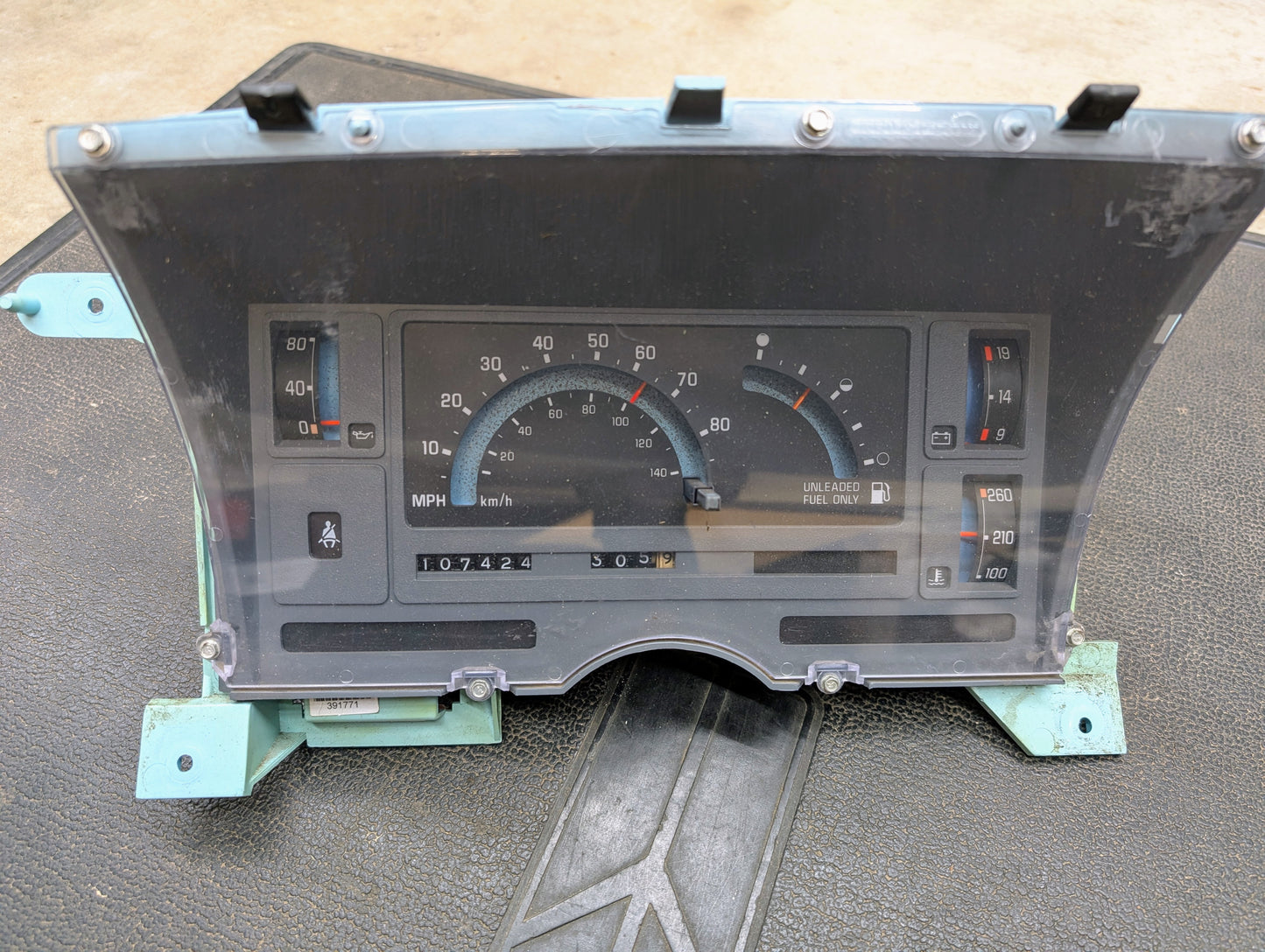 A close-up of the General Motors Low Mile 107K OEM Instrument Cluster for 1986-1994 Chevy S10 Blazer shows analog speed, fuel, temp, and mileage gauges behind a clear cover with turquoise brackets.
