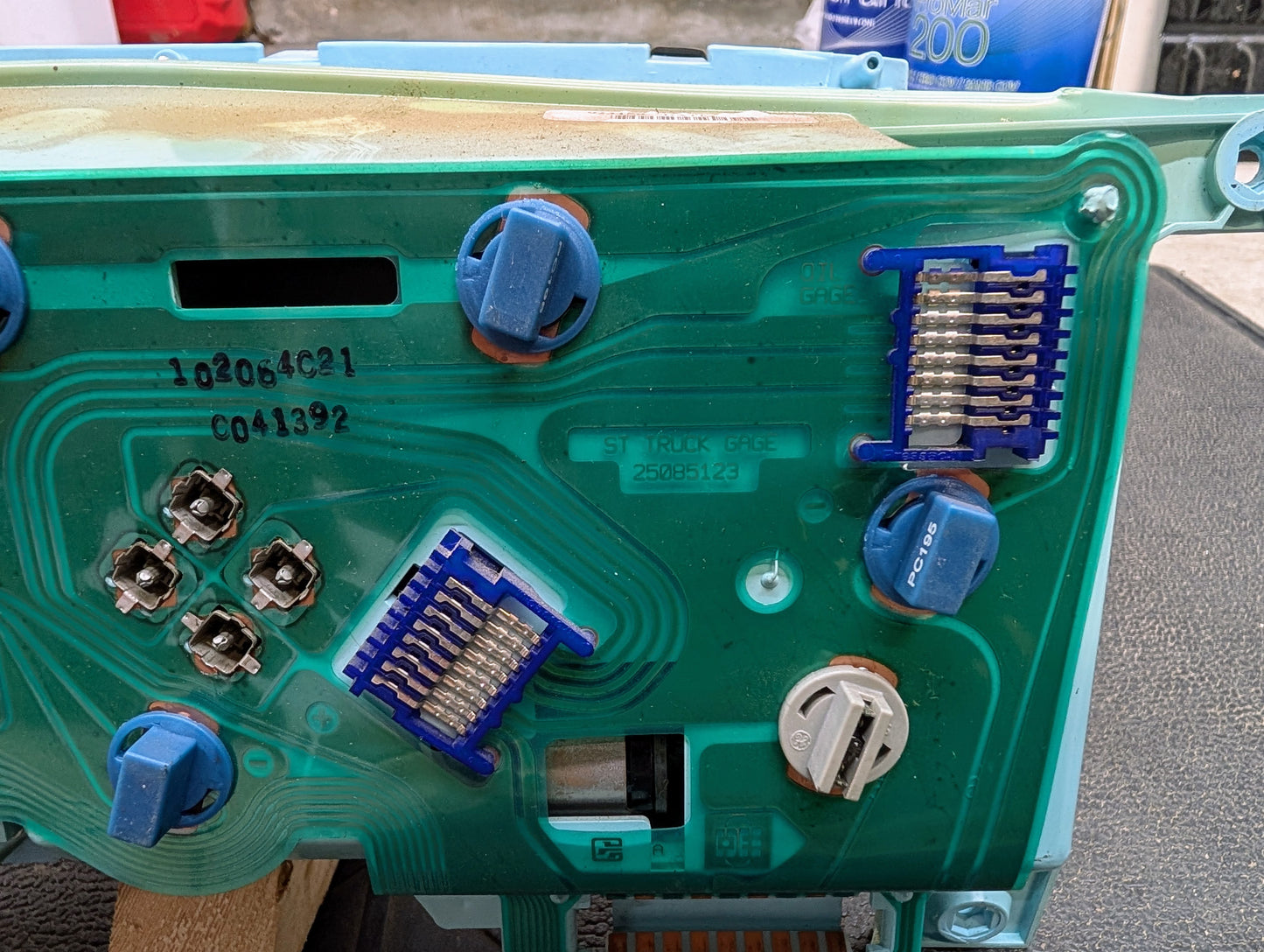 Close-up of a green circuit board from a General Motors Low Mile 107K OEM Instrument Cluster for 1986-1994 Chevy S10 Blazer, showing blue and silver connectors, black numbers like 16142385, and components. A container is blurred in the background.