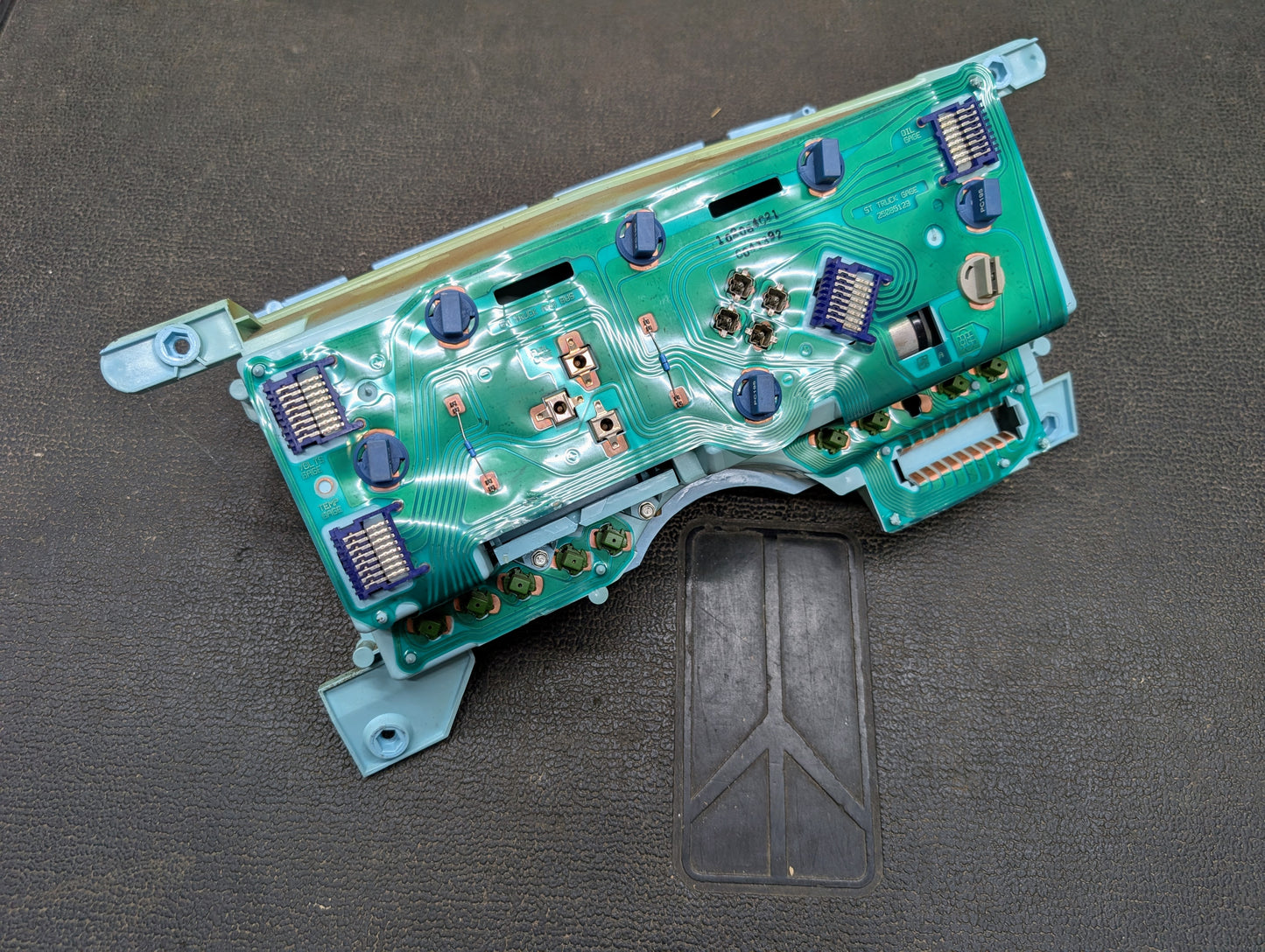 A General Motors Low Mile 107K OEM Instrument Cluster for 1986-1994 Chevy S10 Blazer features a green circuit board with electronic components on a blue plastic frame, shown resting on a textured dark surface.