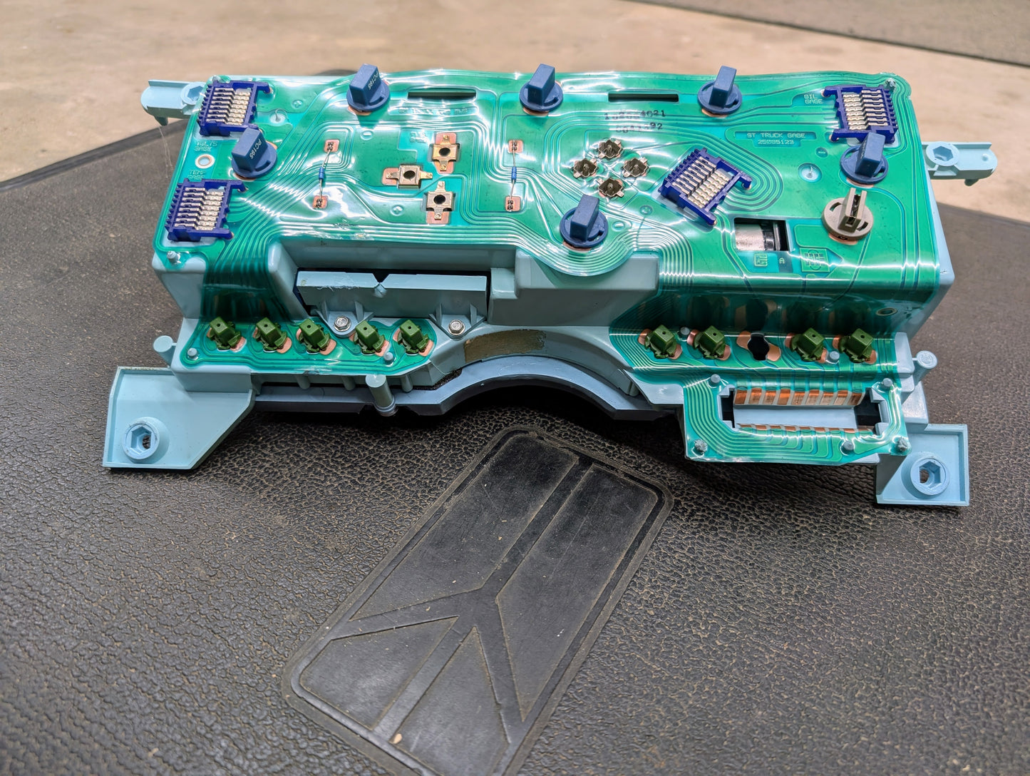 A close-up of a General Motors Low Mile 107K OEM Instrument Cluster for 1986-1994 Chevy S10 Blazer, showing green wiring, blue connectors, and electronic parts on a black surface. Part number: 16142385.