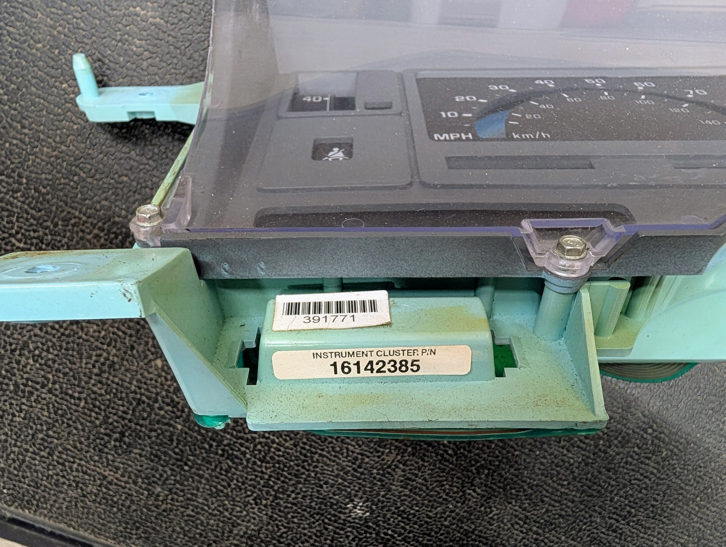 Close-up of a Low Mile 107K OEM Instrument Cluster by General Motors for 1986-1994 Chevy S10 Blazer, showing the barcode sticker, part number 16142385, INSTRUMENT CLUSTER P/N label, green mounting bracket, speedometer, and screws.