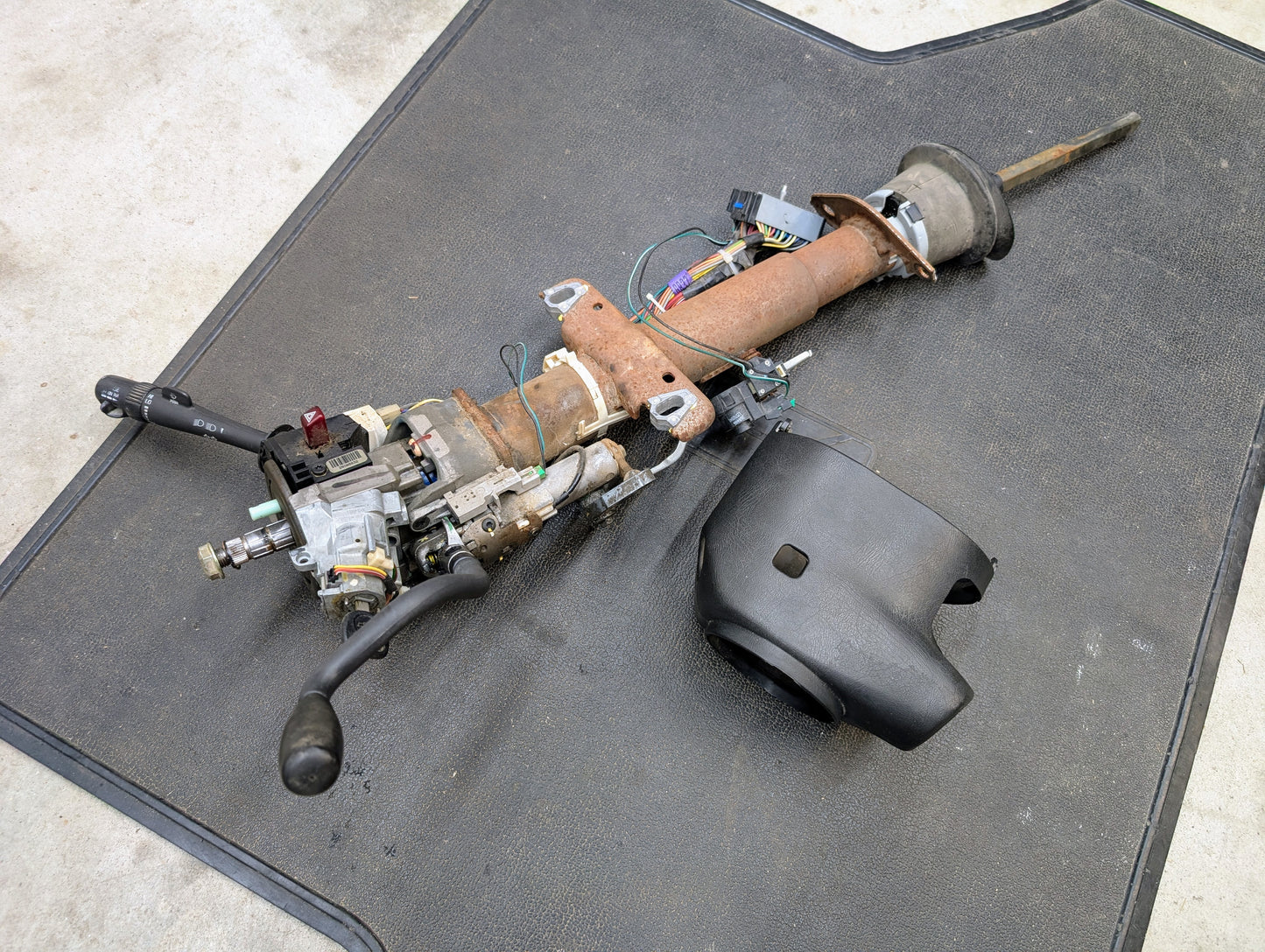 A General Motors OEM Steering Column with tilt, cruise control, and column shift for 1998-2005 Chevy S10, Blazer, and more is shown disassembled with wiring, switches, control stalk, and a black plastic column cover on a black mat.