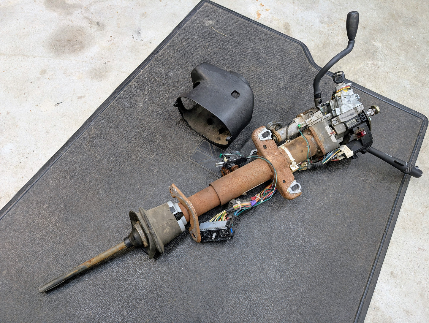A General Motors OEM Steering Column w/Tilt, Cruise Control, and Column Shift for 1998-2005 Chevy S10 Blazer and more lies on a black mat with exposed wiring and a detached black plastic cover.