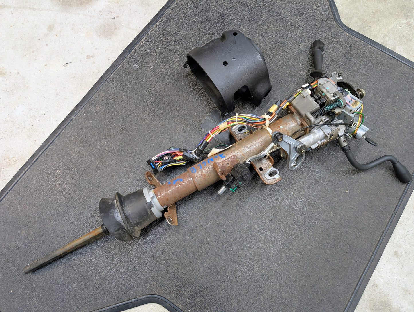 A General Motors OEM Steering Column with tilt, cruise control, and column shift for 1998-2005 Chevy S10 Blazer and more lies on a black mat, wiring harnesses and components attached, viewed from above.