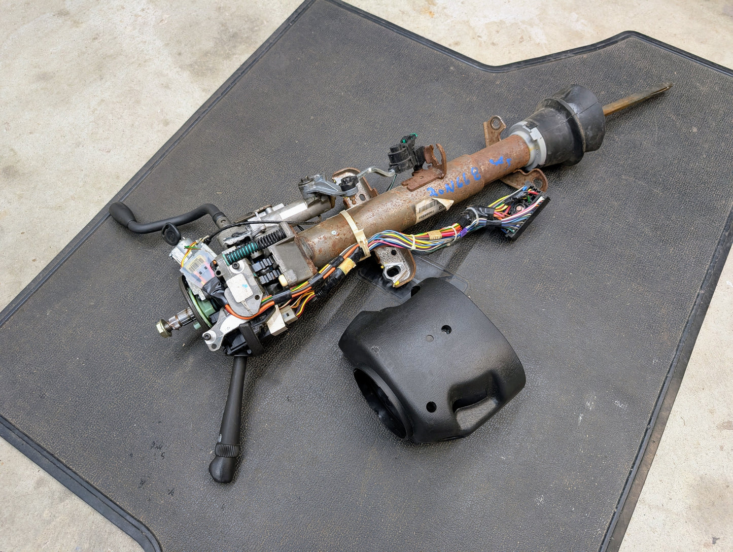 A General Motors OEM Steering Column w/Tilt, Cruise Control, and Column Shift for 1998-2005 Chevy S10 Blazer and more sits on a black mat with visible wires, control levers, and a detached black plastic cover on a concrete floor.