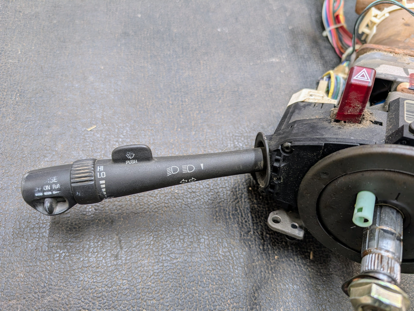 A detailed view of the General Motors OEM Steering Column w/Tilt, Cruise Control, and Column Shift for 1998-2005 Chevy S10 Blazer shows its turn signal and headlight control lever with wiring and dashboard parts on a textured black surface.