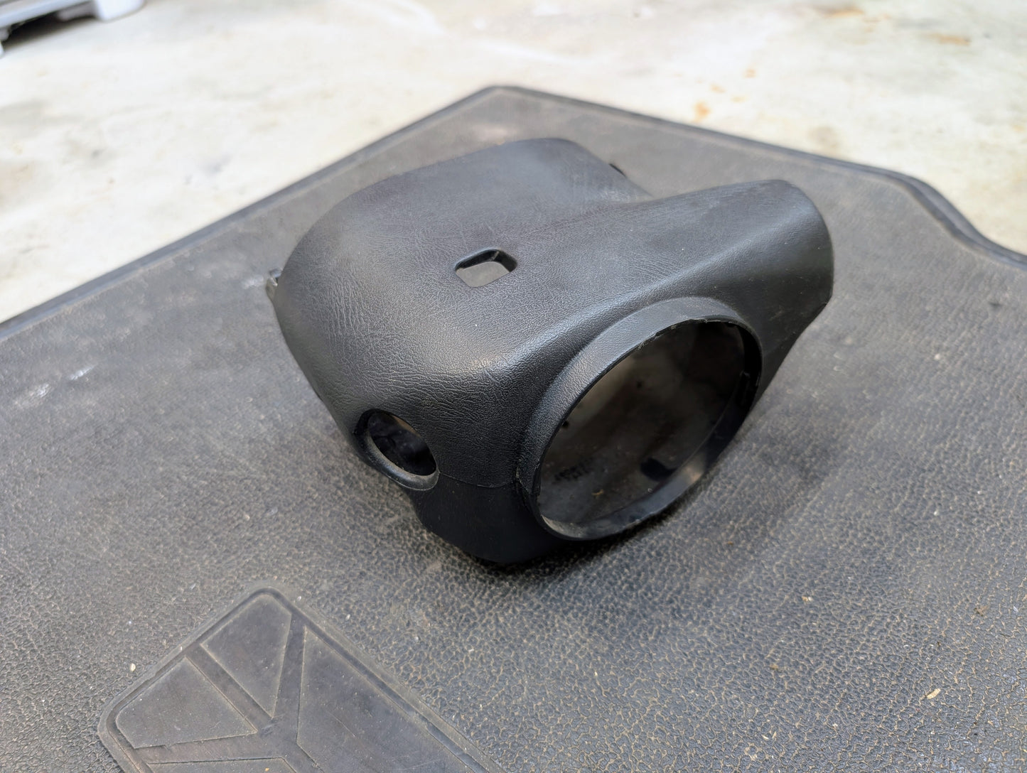 A black General Motors OEM steering column cover for 1998-2005 Chevy S10 Blazer with tilt, cruise control, and column shift sits on a textured floor mat against a concrete background.