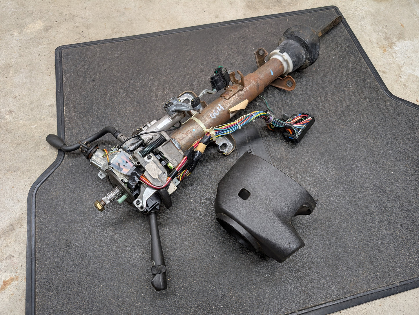 A General Motors OEM Steering Column w/Tilt, Cruise Control, and Column Shift for 1998-2005 Chevy S10 Blazer and more is shown disassembled with wiring, levers, and a black cover on a black mat on a concrete floor.