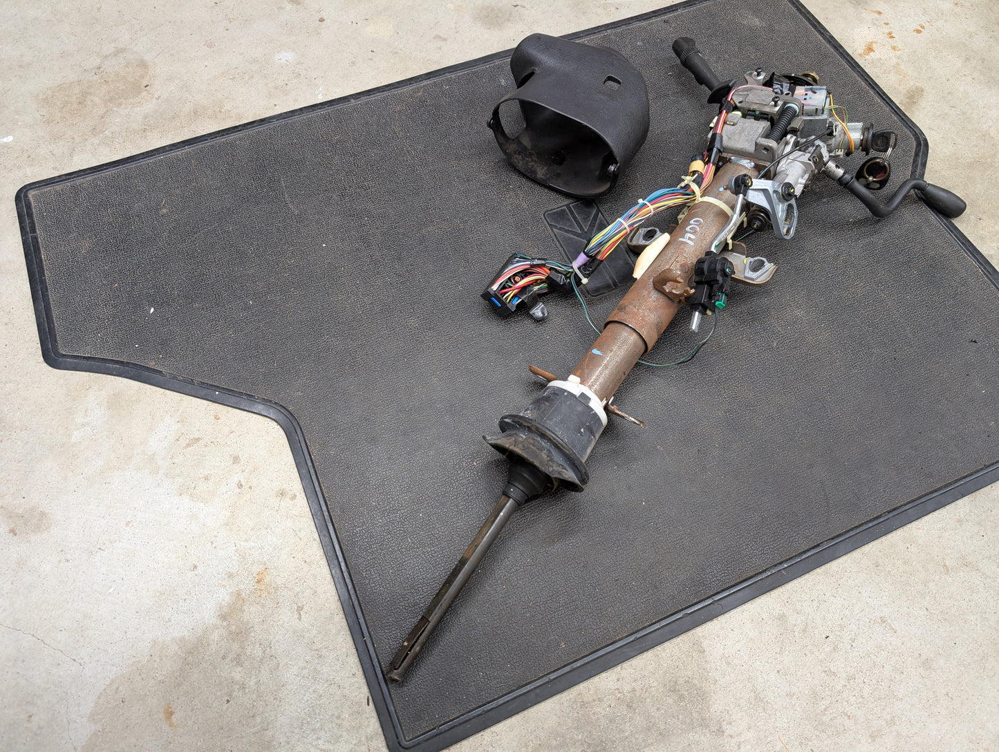 A General Motors OEM Steering Column w/Tilt, Cruise Control, and Column Shift for 1998-2005 Chevy S10/Blazer rests on a black floor mat with exposed wiring and components; the plastic cover is removed and lies nearby.