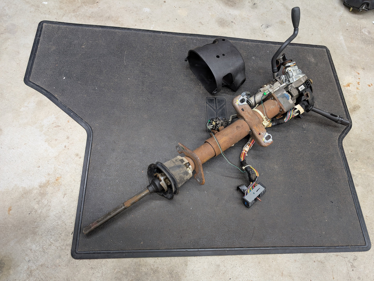 A General Motors OEM Steering Column w/Tilt, Cruise Control, and Column Shift for 1998-2005 Chevy S10 Blazer and more sits on a black mat on a concrete floor, with wiring, switches, and levers attached.