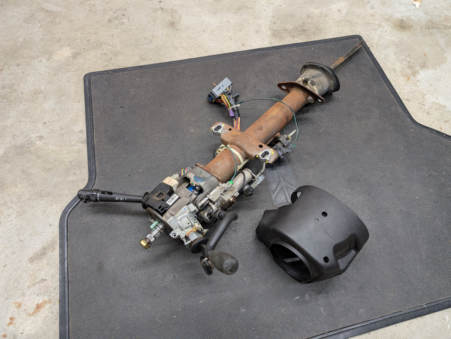 A General Motors OEM Steering Column w/Tilt, Cruise Control, and Column Shift for 1998-2005 Chevy S10 Blazer rests on a black mat next to a separate plastic cover on a concrete floor.