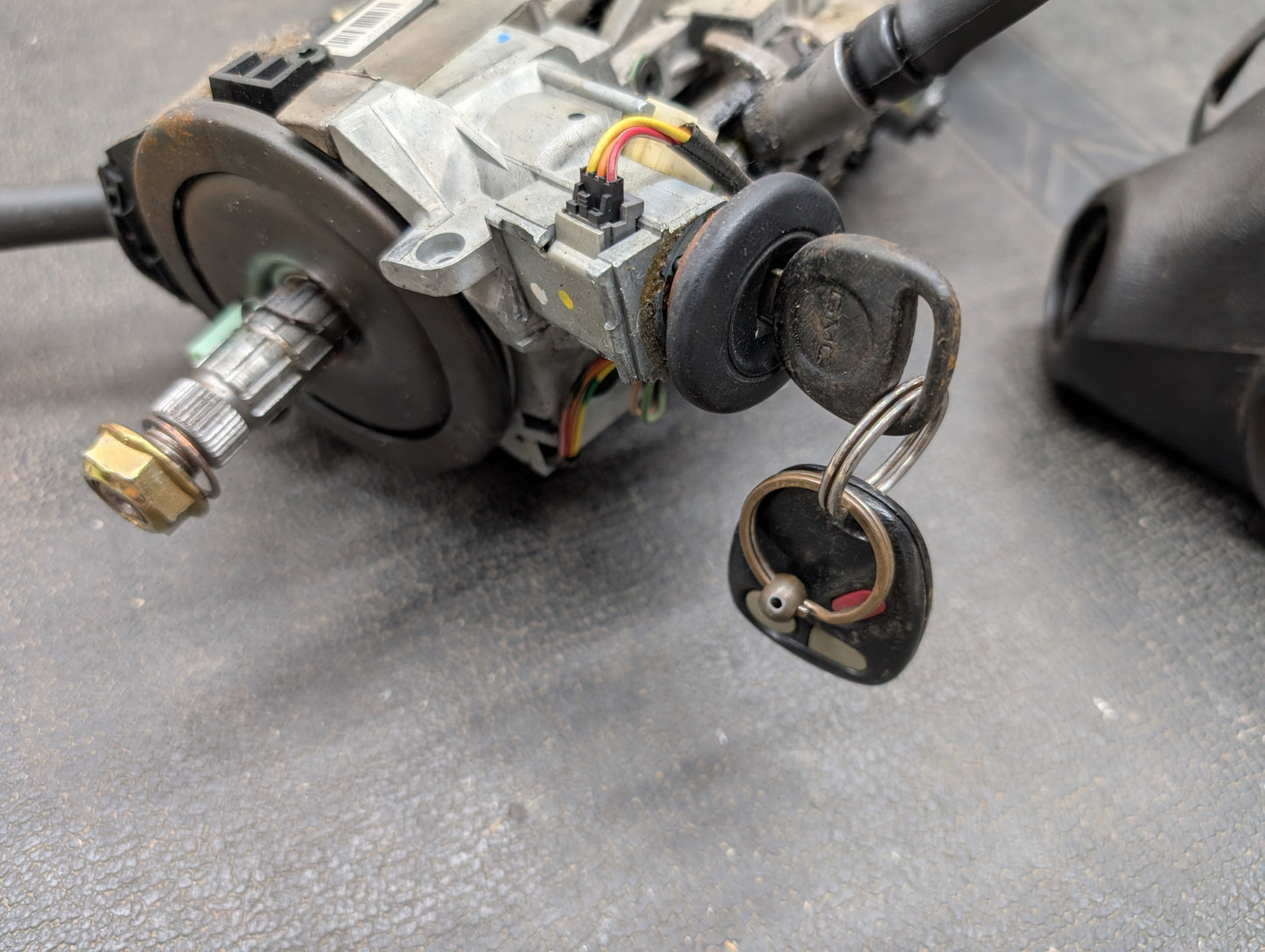 A close-up of a General Motors OEM Steering Column with tilt, cruise control, and column shift for 1998-2005 Chevy S10 Blazer, featuring an ignition switch assembly with attached wires and a key with a black plastic head.