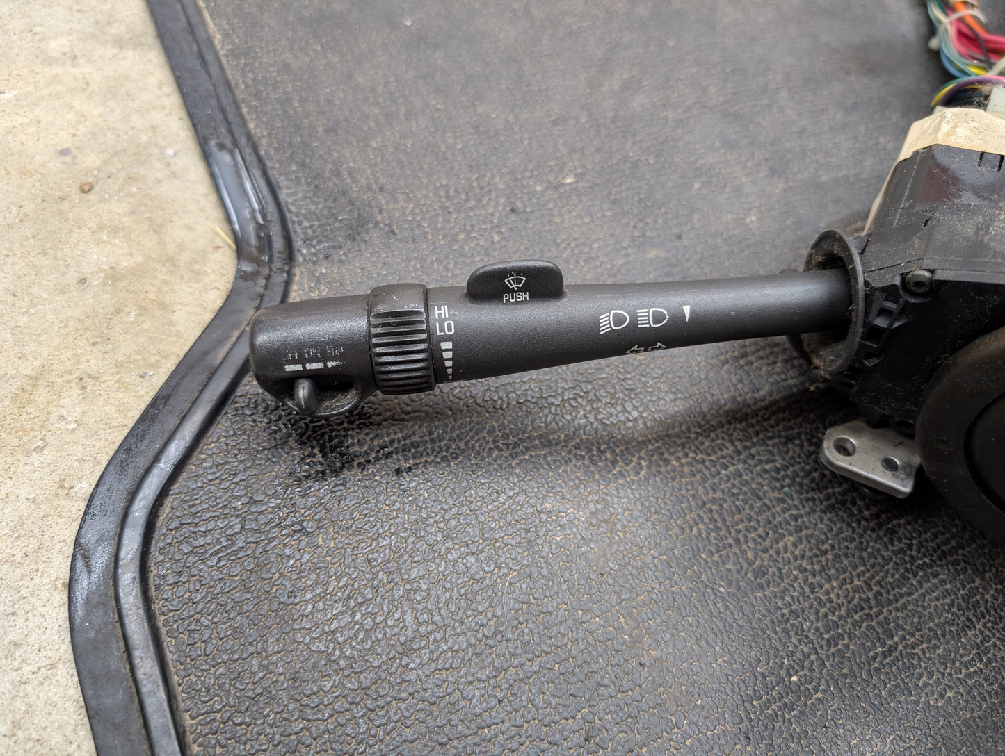 A close-up of a General Motors OEM Steering Column w/Tilt, Cruise Control, and Column Shift for 1998-2005 Chevy S10 Blazer shows the headlight and wiper control stalk with various symbols, plus multicolored background wires.