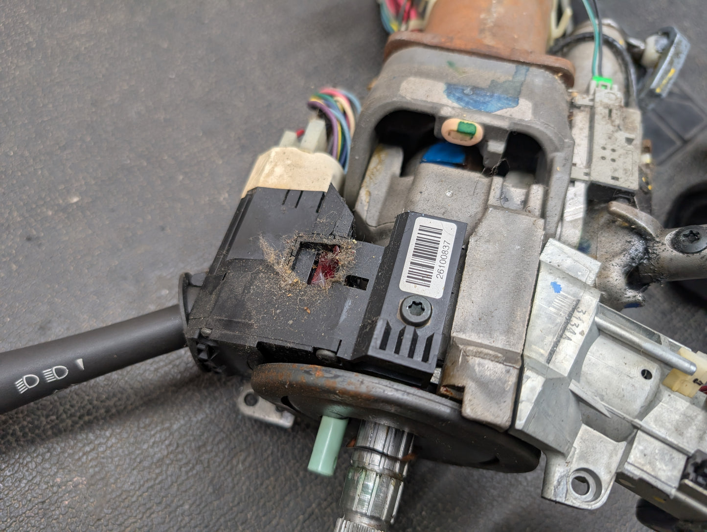 A close-up of a worn General Motors OEM Steering Column with tilt, cruise control, and column shift for 1998-2005 Chevy S10 Blazer, showing wiring, barcode label, and turn signal lever on a dark textured surface.
