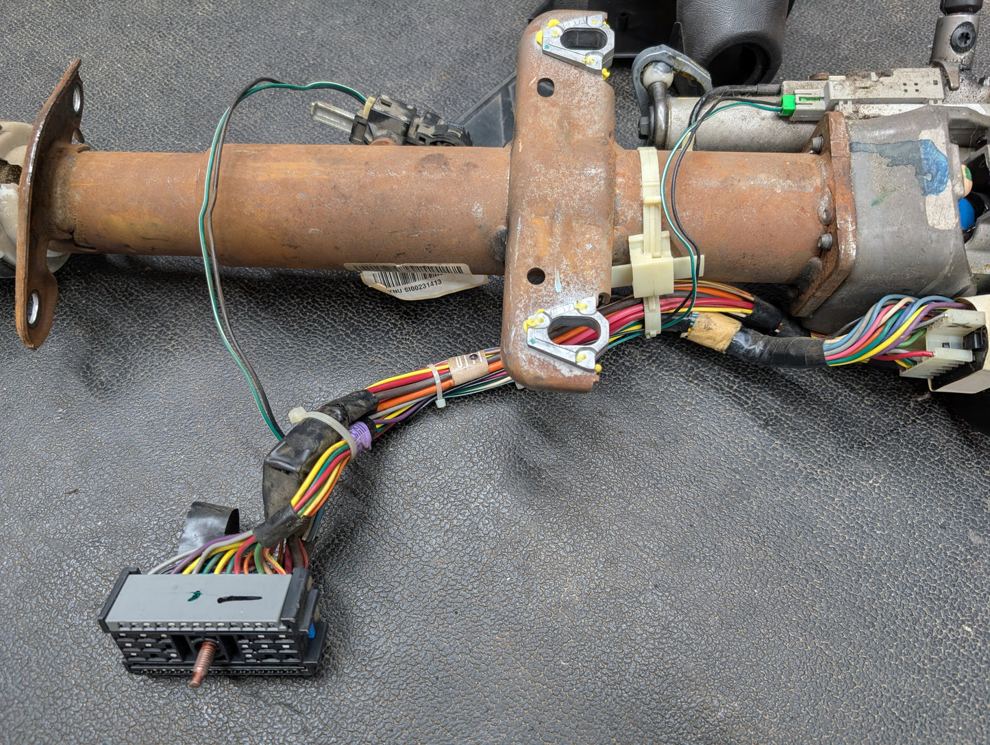 A General Motors OEM steering column with tilt, cruise control, and column shift for 1998-2005 Chevy S10/Blazer rests on a textured black surface, showing its wiring harnesses, colorful wires, and electrical connectors.