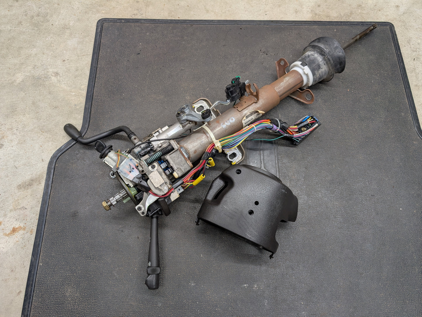 A General Motors OEM Steering Column w/Tilt, Cruise Control, and Column Shift for 1998-2005 Chevy S10 Blazer rests on a black mat, with exposed wiring and levers; a black plastic cover lies nearby—fits automatic transmission setups.