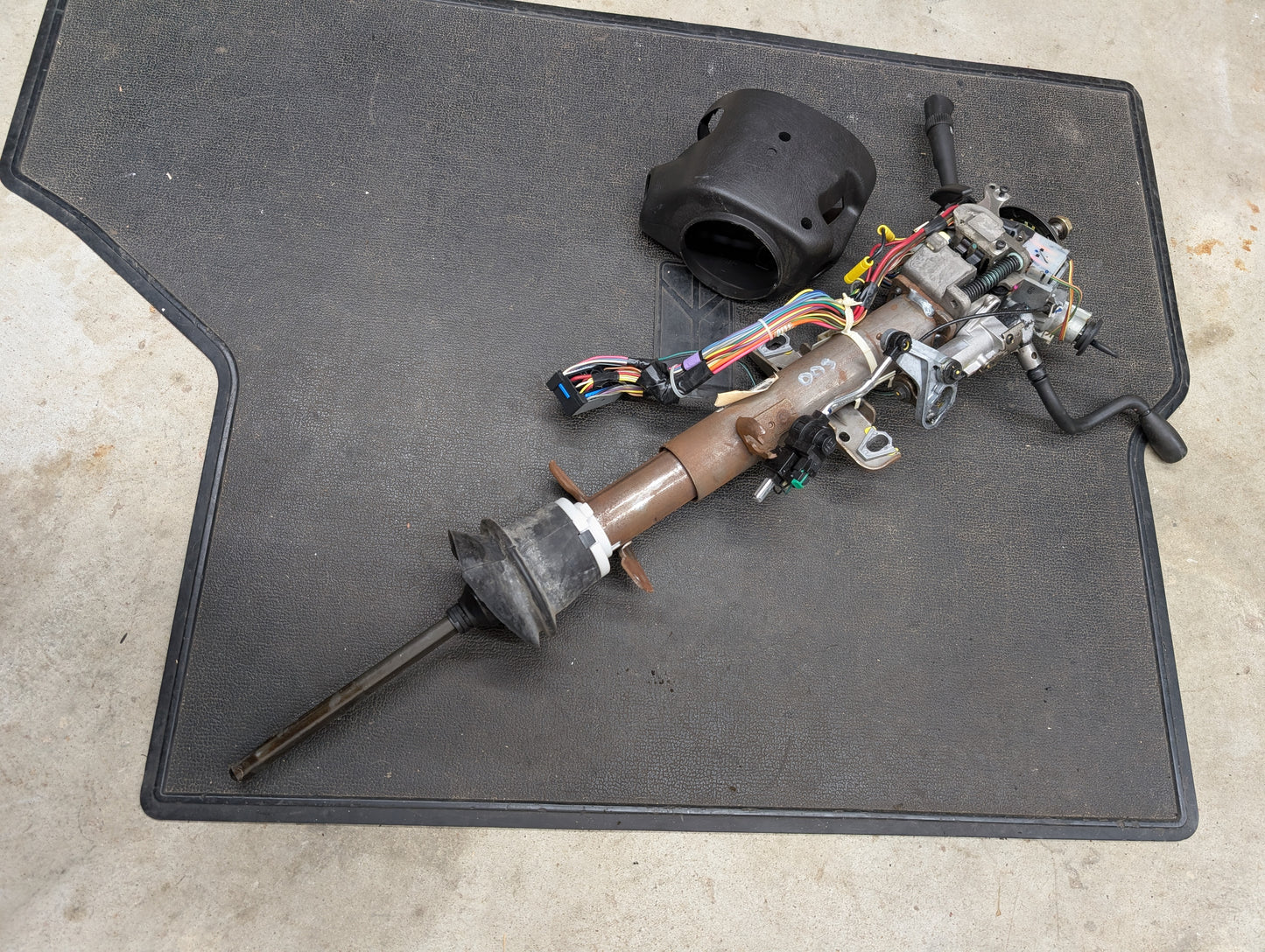 A General Motors OEM Steering Column with tilt, cruise control, and column shift for 1998-2005 Chevy S10 Blazer rests disassembled on a black floor mat with wiring attached; a black plastic cover is nearby.