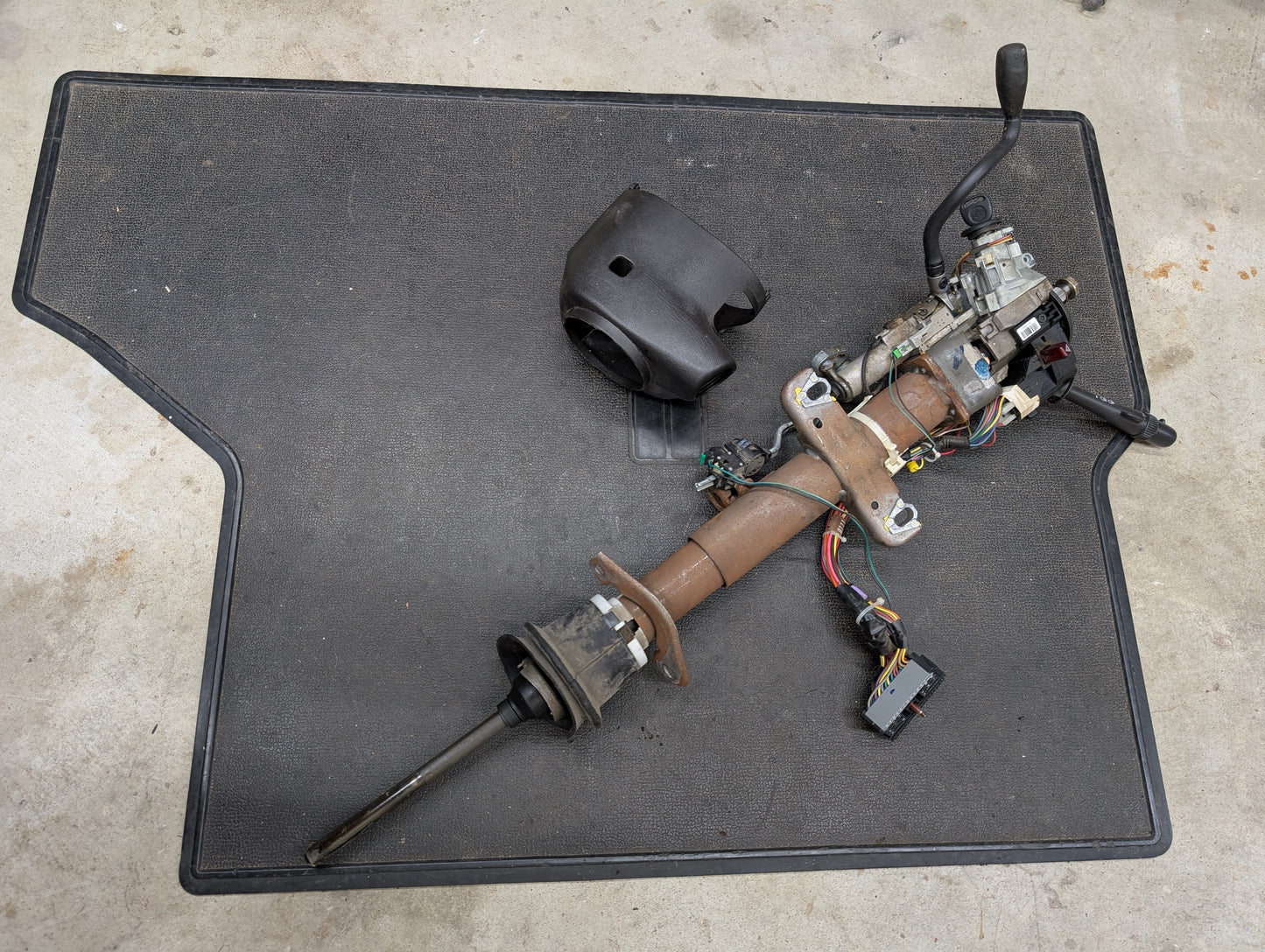 A disassembled General Motors OEM Steering Column w/Tilt, Cruise Control, and Column Shift for 1998-2005 Chevy S10/Blazer rests with wiring and ignition parts on a mat; a plastic cover for Blazer automatic transmissions is nearby.