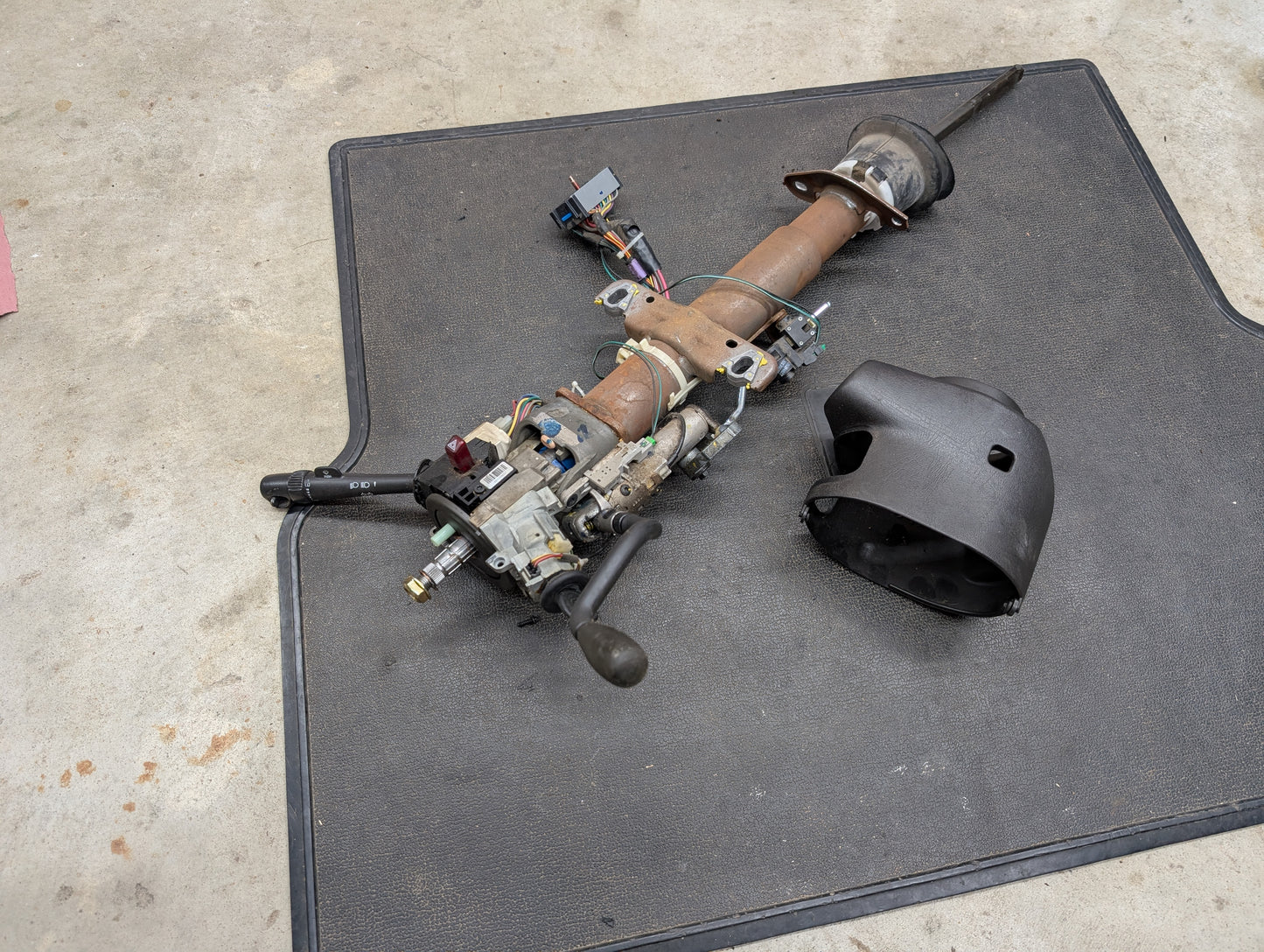 A disassembled General Motors OEM Steering Column w/Tilt, Cruise Control, and Column Shift for 1998-2005 Chevy S10 Blazer lies on a black mat, with exposed wiring and a black plastic cover nearby on the floor.