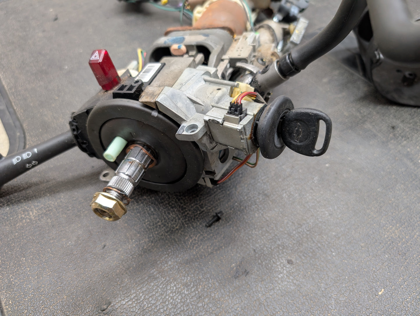 A close-up of a General Motors OEM Steering Column w/Tilt, Cruise Control, and Column Shift for 1998-2005 Chevy S10 Blazer shows the ignition assembly with key, wiring, and connectors on a textured black surface.