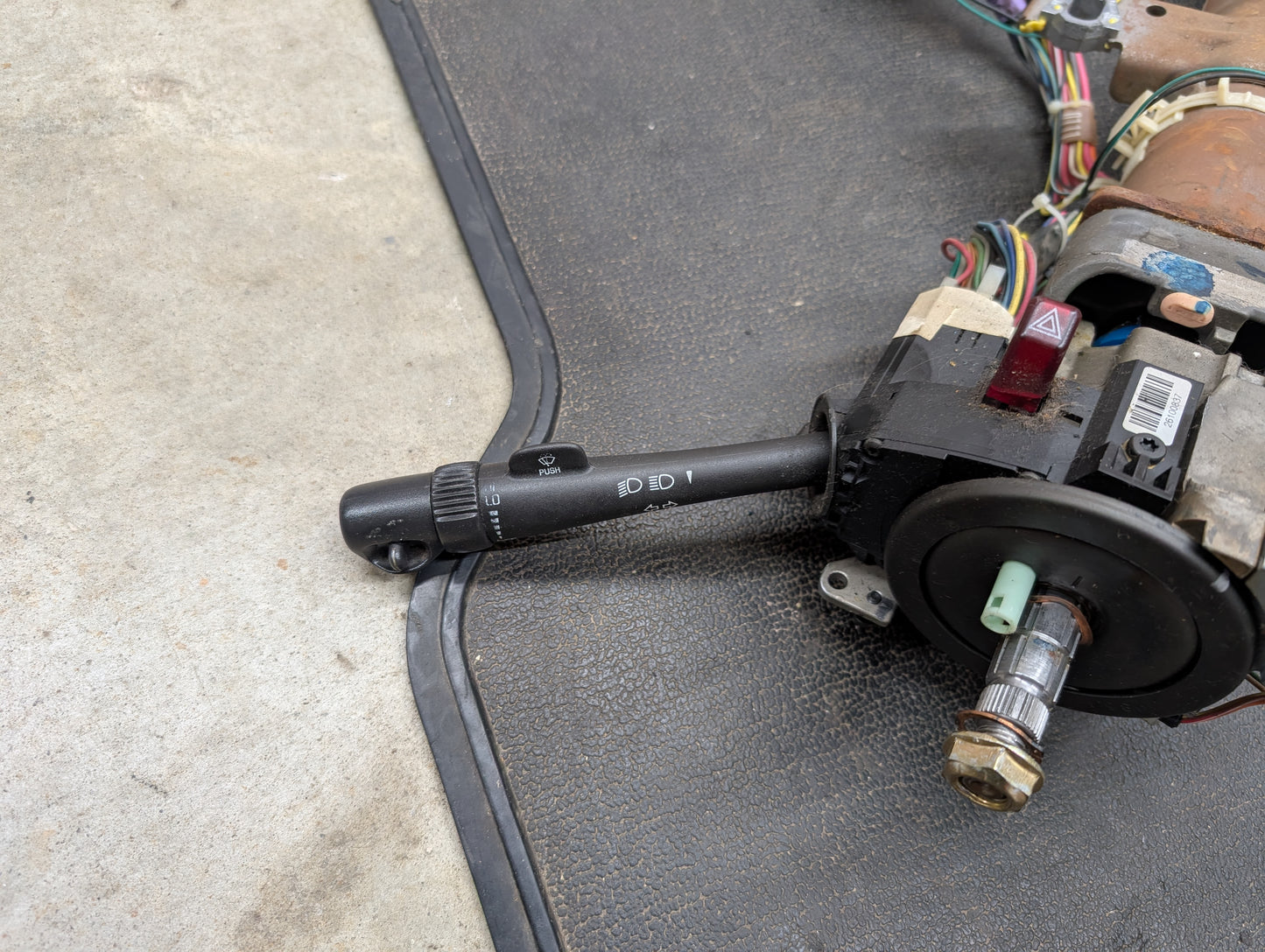 A General Motors OEM Steering Column w/Tilt, Cruise Control, and Column Shift for 1998-2005 Chevy S10 Blazer is shown partially disassembled with exposed wires on a black mat atop a concrete floor.
