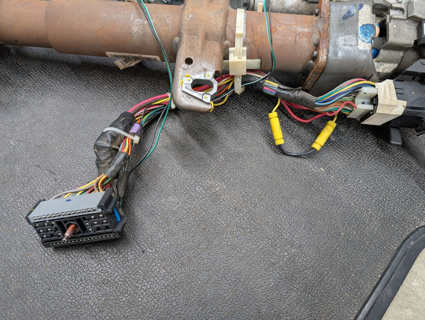 A close-up of a General Motors OEM Steering Column w/Tilt, Cruise Control, and Column Shift for 1998-2005 Chevy S10 Blazer, shows multiple exposed wires and connectors on a textured black surface; some connectors are unplugged.